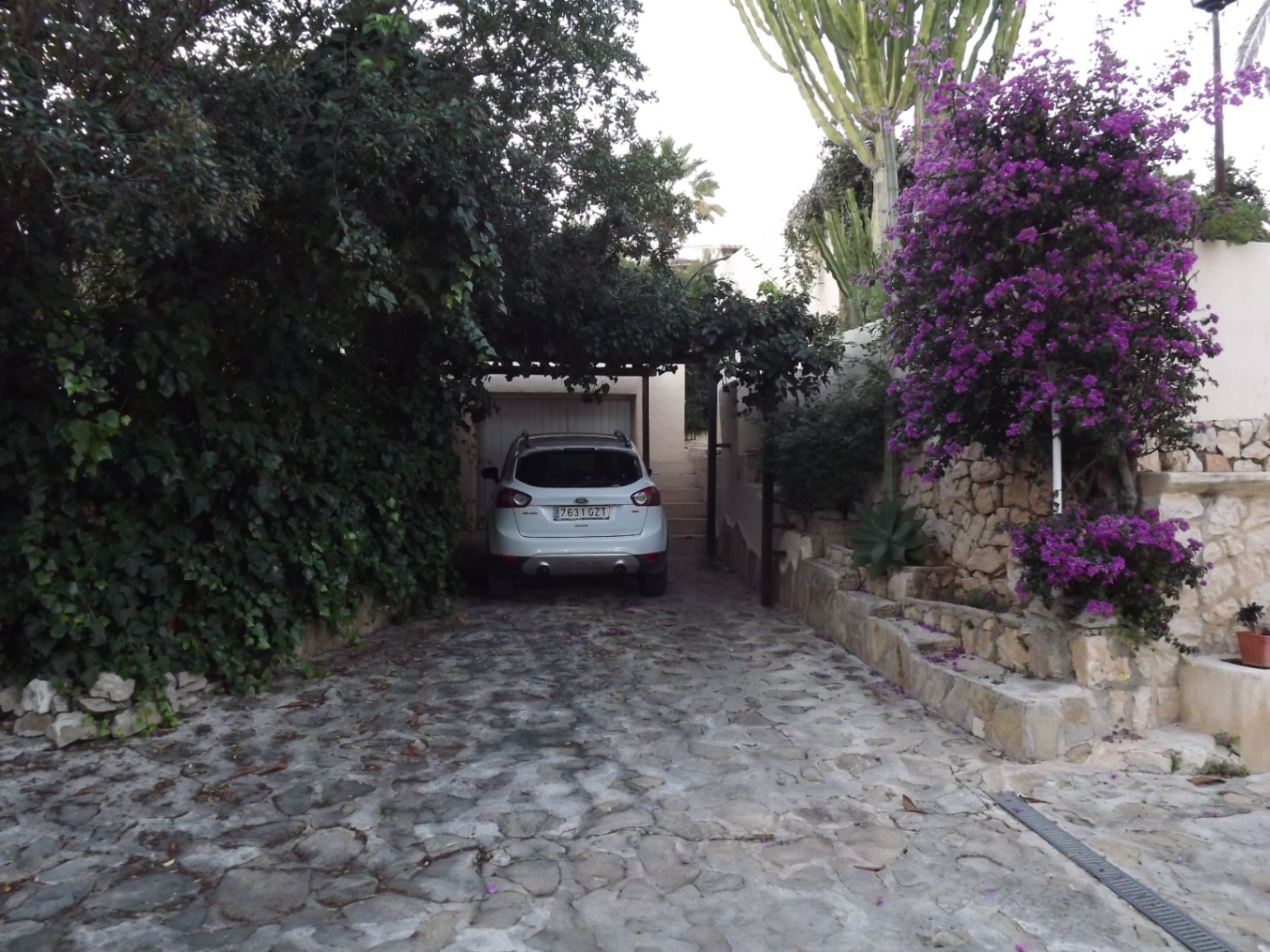 Natursteingepflasterte Einfahrt mit bewachsenem Carport, Trockensteinmauern und blühenden Bougainvilleen, die einen charmanten Zugang bieten.