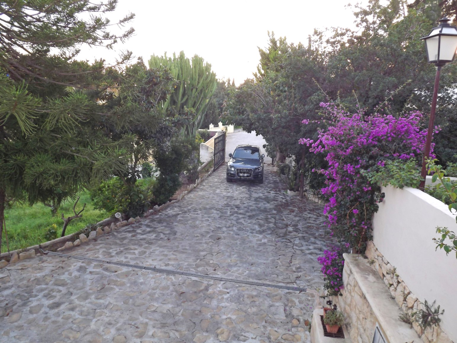 Privatweg mit Natursteinpflasterung, umgeben von mediterraner Bepflanzung, Bougainvillea und einer Umfassungsmauer aus weißem Putz und Naturstein.