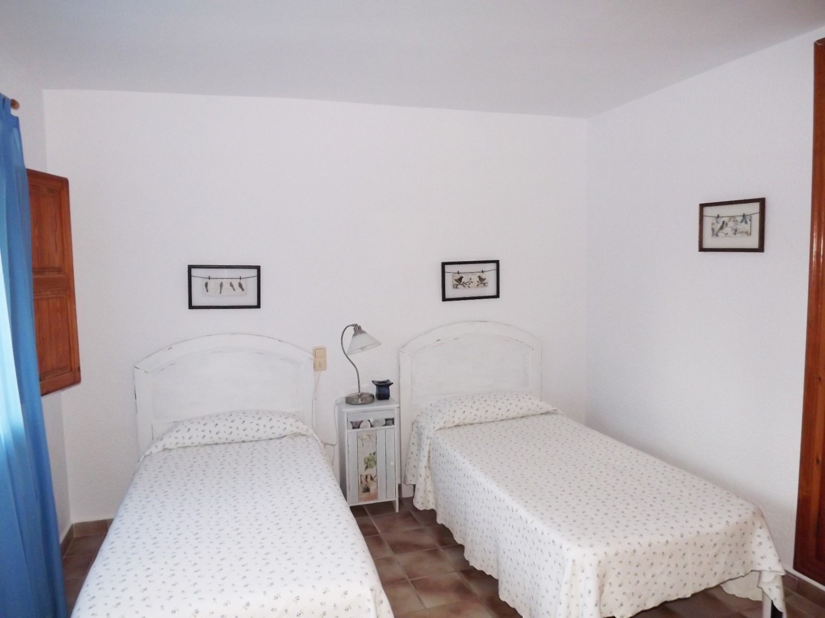 Schlafzimmer mit zwei Einzelbetten, weißen Kopfteilen und rustikalen Terrakotta-Fliesen. Mit traditionellen Holzfensterläden und hellem, natürlichem Licht.