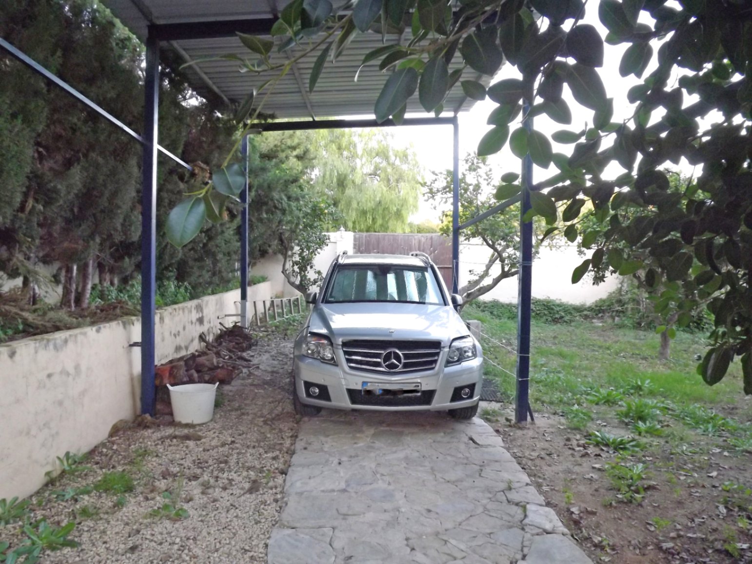 Überdachter Parkplatz mit Metallkonstruktion und Natursteinpflasterung, eingebettet in eine grüne Umgebung mit altem Baumbestand.