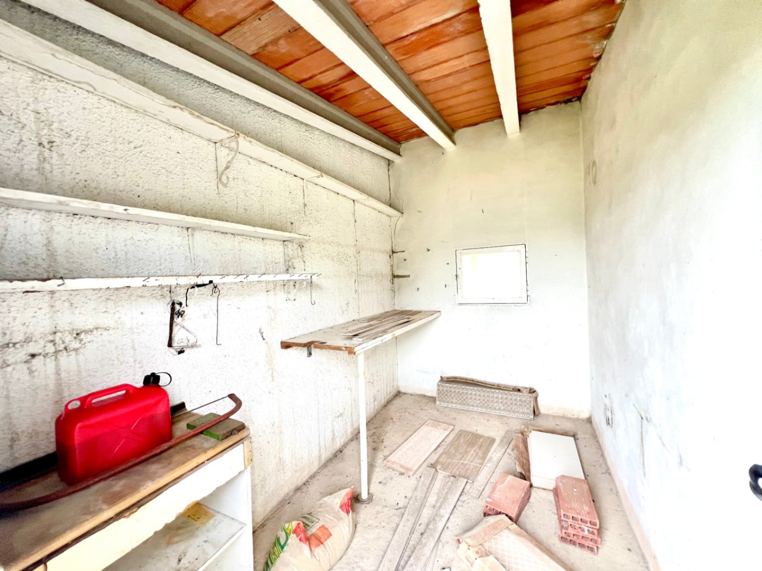 Funktionaler Abstellraum mit sichtbaren Holzbalken und Terrakotta-Decke. Ausgestattet mit Regalen, einer Werkbank und natürlichem Licht durch ein kleines Fenster.