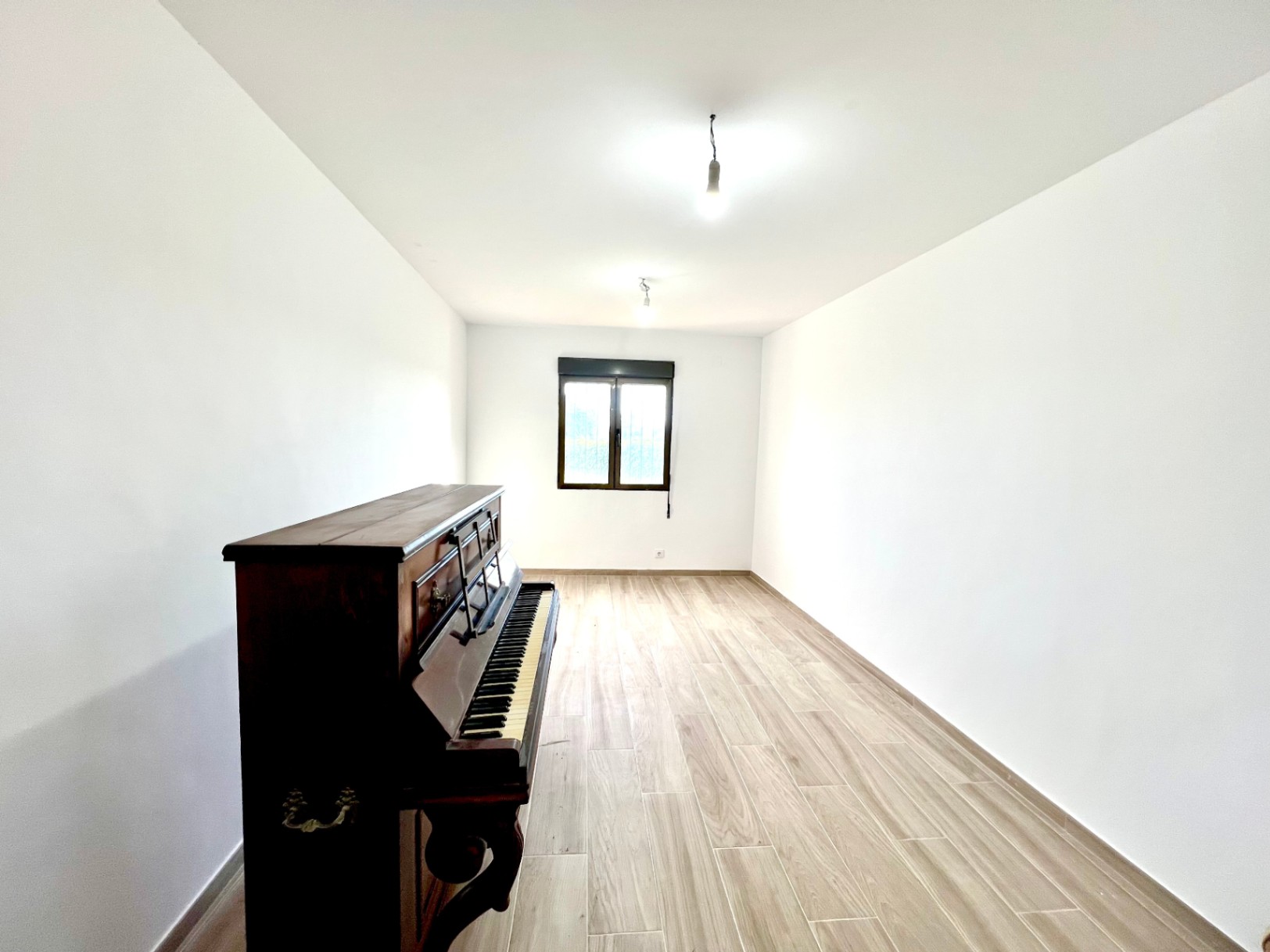Heller, länglicher Raum mit Bodenfliesen in Holzoptik, weißen Wänden und einem klassischen Klavier. Ein Fenster mit dunklem Rahmen sorgt für Tageslicht.