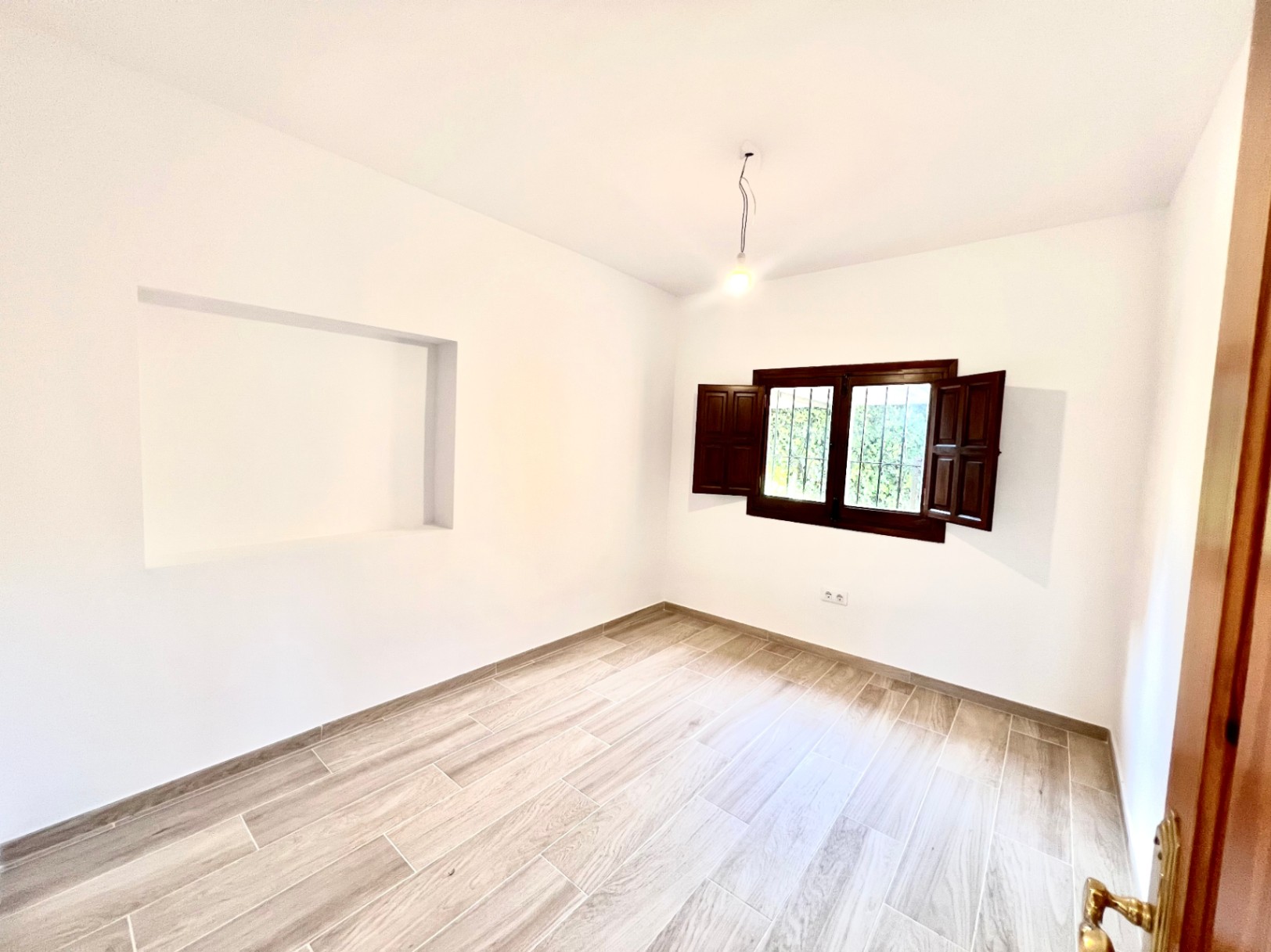 Helles Schlafzimmer mit Bodenfliesen in Holzoptik, einer Wandnische und einem Fenster mit traditionellen dunklen Holzklappläden.