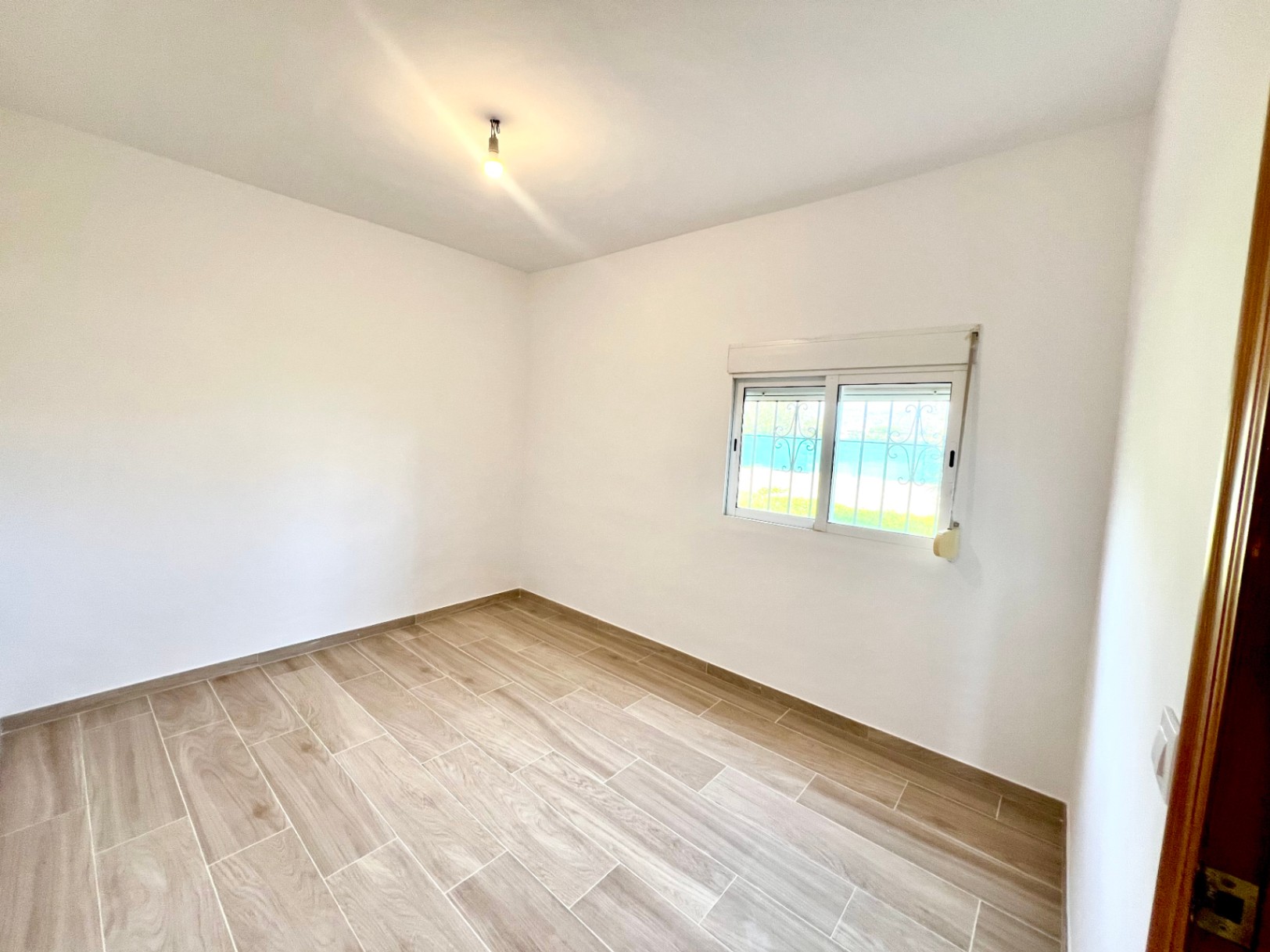 Helles Schlafzimmer mit keramischen Fliesen in Holzoptik, glatten weißen Wänden und Fenster mit Sicherheitsgitter und integriertem Rollladen.