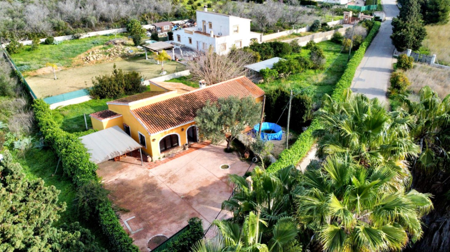 Luftaufnahme der freistehenden Villa mit gelber Fassade, Ziegeldach, großem gepflastertem Innenhof, überdachtem Carport und weitläufigem Naturgrundstück.