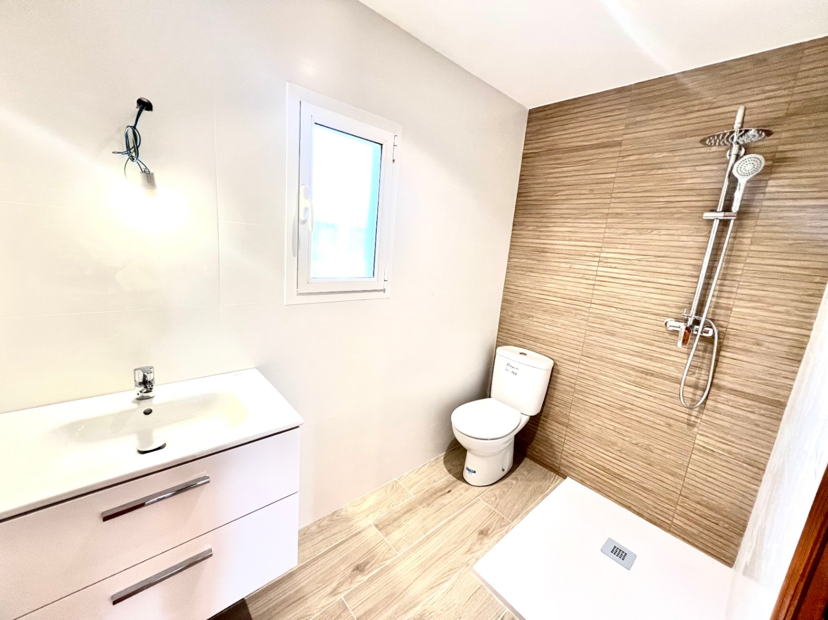 Modernes Badezimmer mit begehbarer Dusche, Fliesen in Holzoptik und weißem Waschtisch. Ein PVC-Fenster sorgt für natürliches Licht und Belüftung.