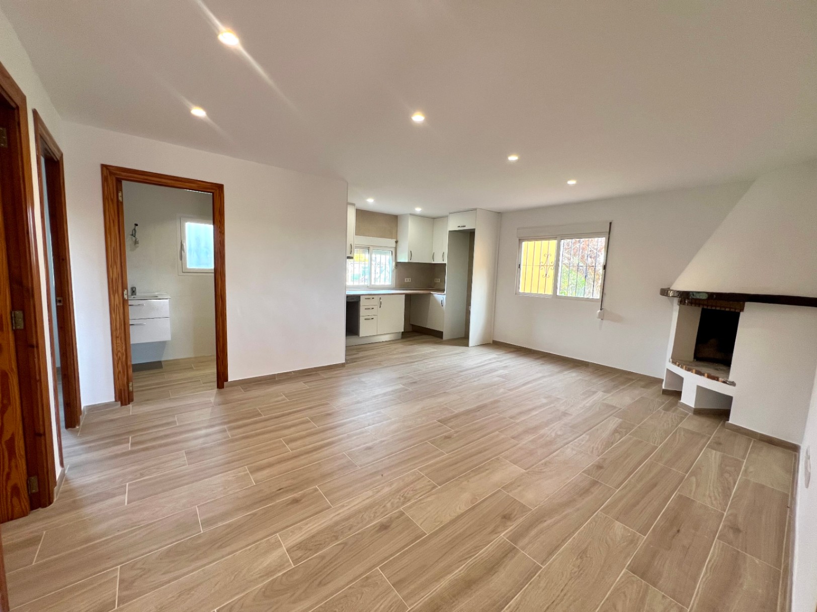 Offener Wohnbereich mit Eckkamin, integrierter weißer Küche und Keramikböden in Holzoptik unter modernen Deckeneinbaustrahlern.