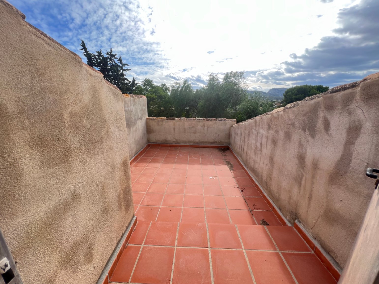 Dachterrasse im Obergeschoss mit Terrakotta-Fliesen und hohen verputzten Mauern, die einen weiten Blick auf die umliegenden Bäume und Berge bietet.