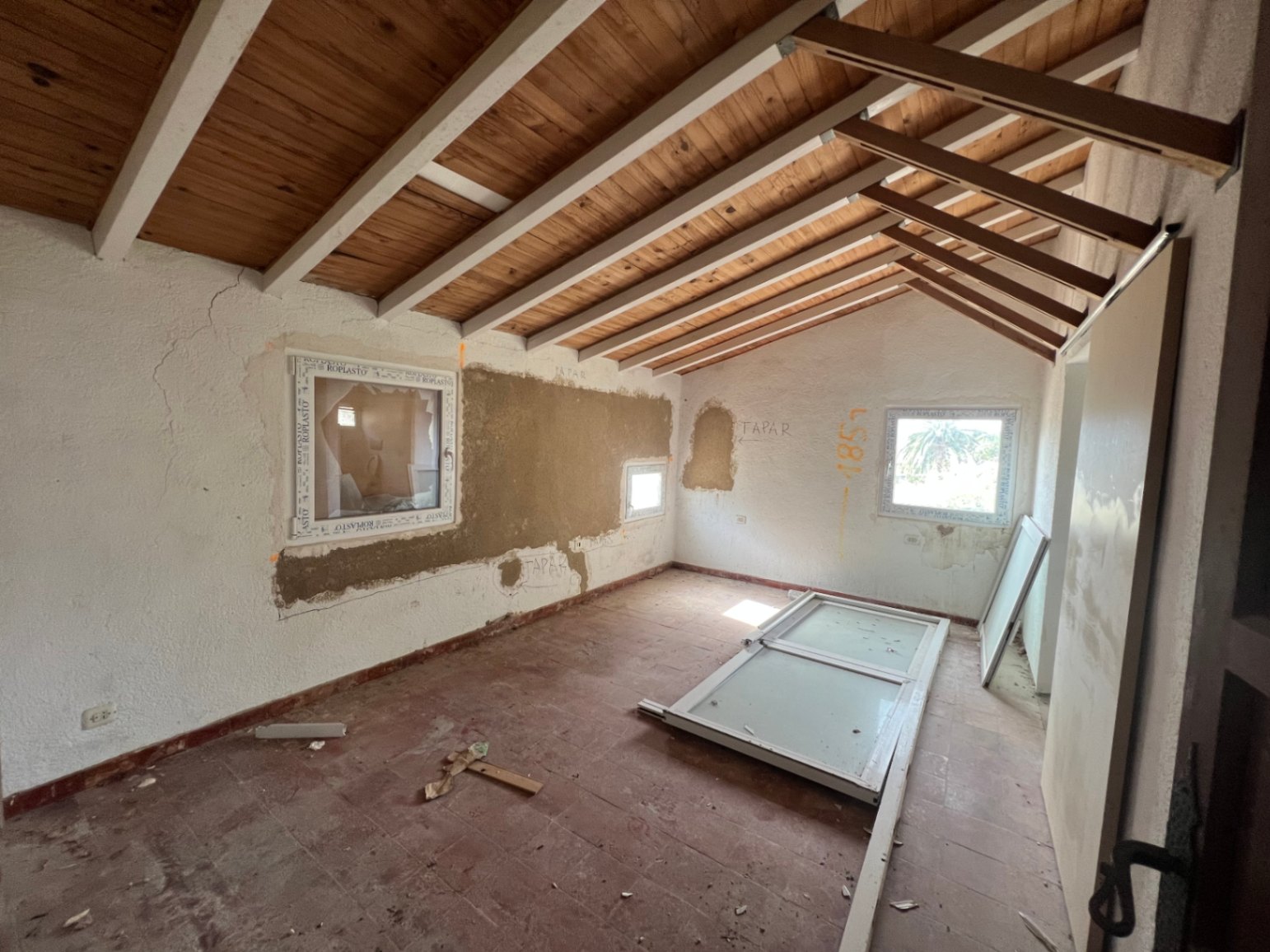 Innenraum in Renovierung mit sichtbaren Holzbalkendecken, Terrakottaböden und neu installierten PVC-Fensterrahmen für verbesserte Isolierung.
