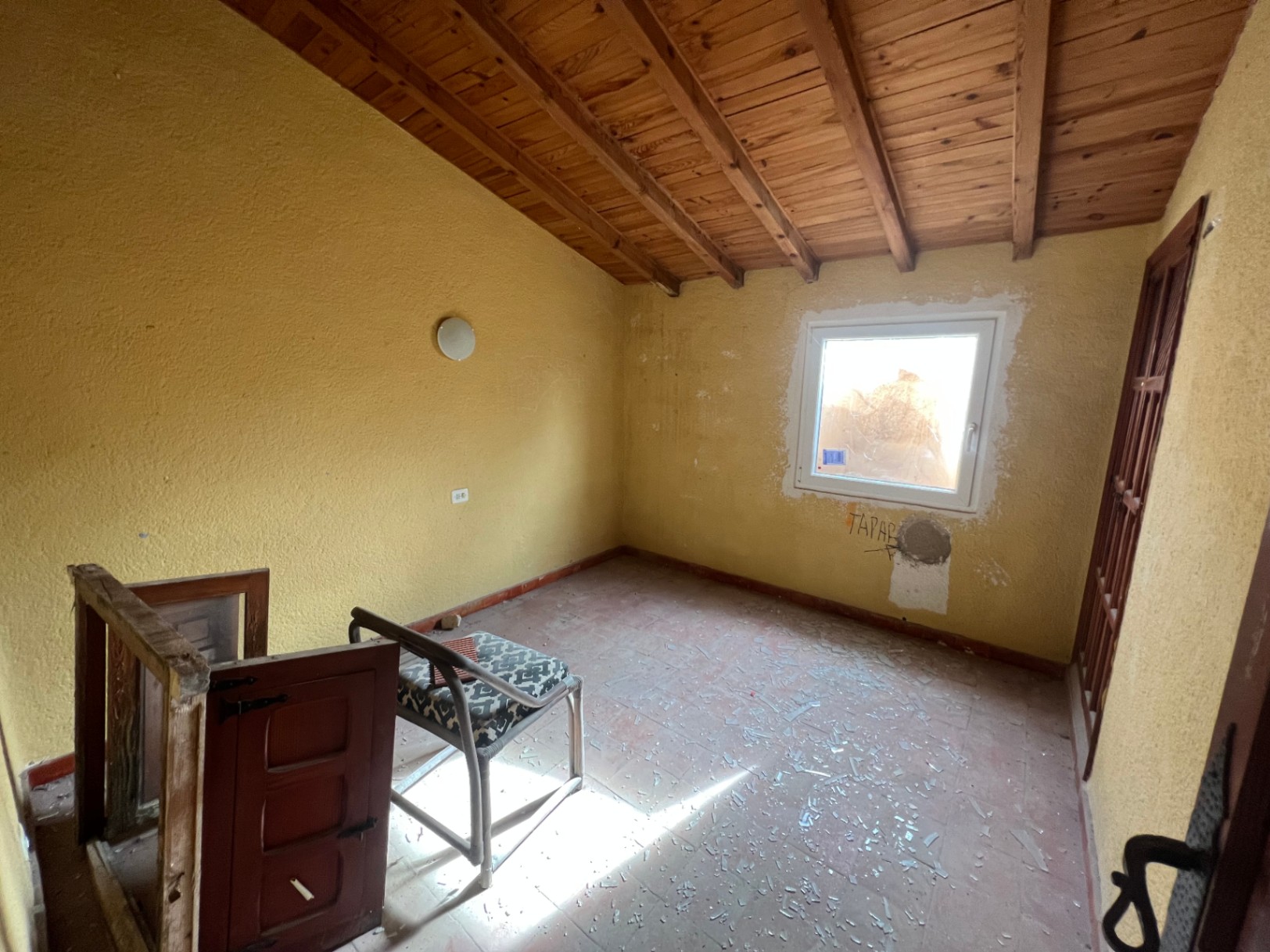 Innenraum mit schräger Holzbalkendecke, Terrakottaboden und gelben Wänden. Der Raum ist renovierungsbedürftig und verfügt über ein Fenster für natürliches Licht.