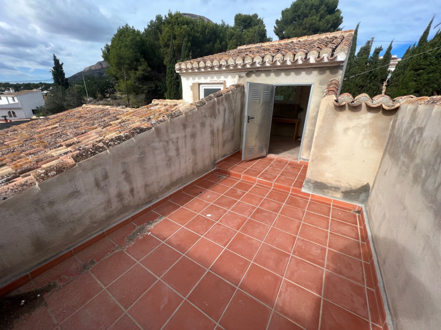 Obere Terrasse mit roten Keramikfliesen und gemauerten Brüstungen, die Zugang zu einem separaten Studio unter einem traditionellen Ziegeldach bietet.