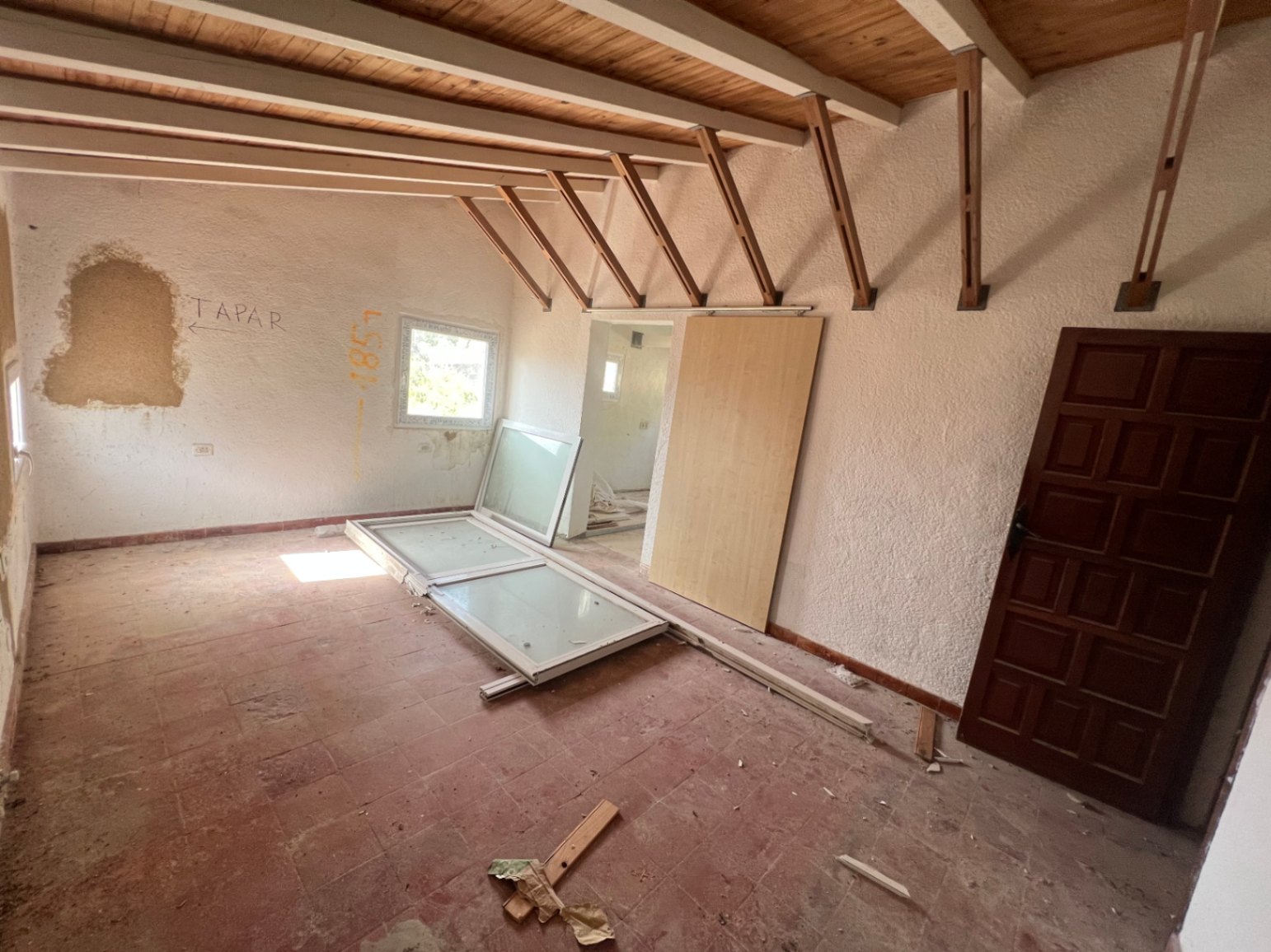 Renovierungsbedürftiger Innenraum mit sichtbaren Deckenbalken aus Holz, originalen Terrakottaböden und zur Montage bereitstehenden Fensterrahmen.
