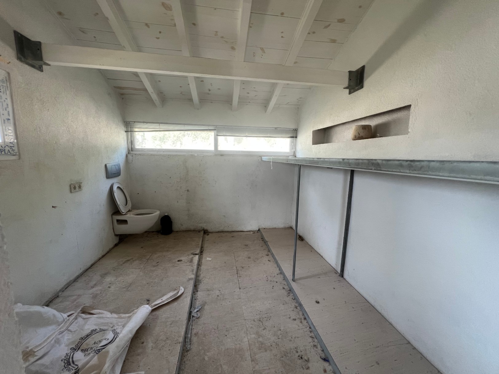 Renovierungsbedürftiger Innenraum mit weißen Holzbalken, horizontalem Fenster, WC und langen Regalen. Vielseitig nutzbar als Studio oder Lagerraum.