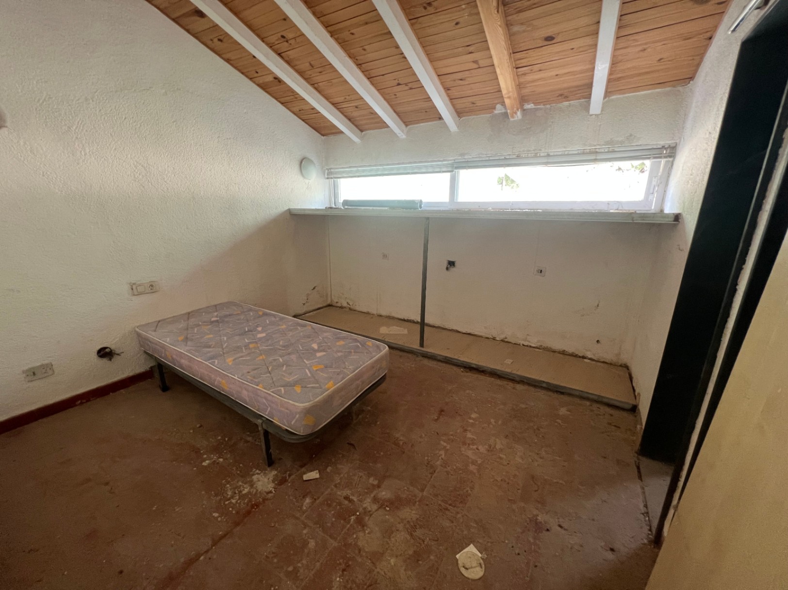 Schlafzimmer mit Dachschräge, sichtbaren Holzbalken und rustikalem Fliesenboden, das ein erhebliches Potenzial für eine umfassende Renovierung bietet.