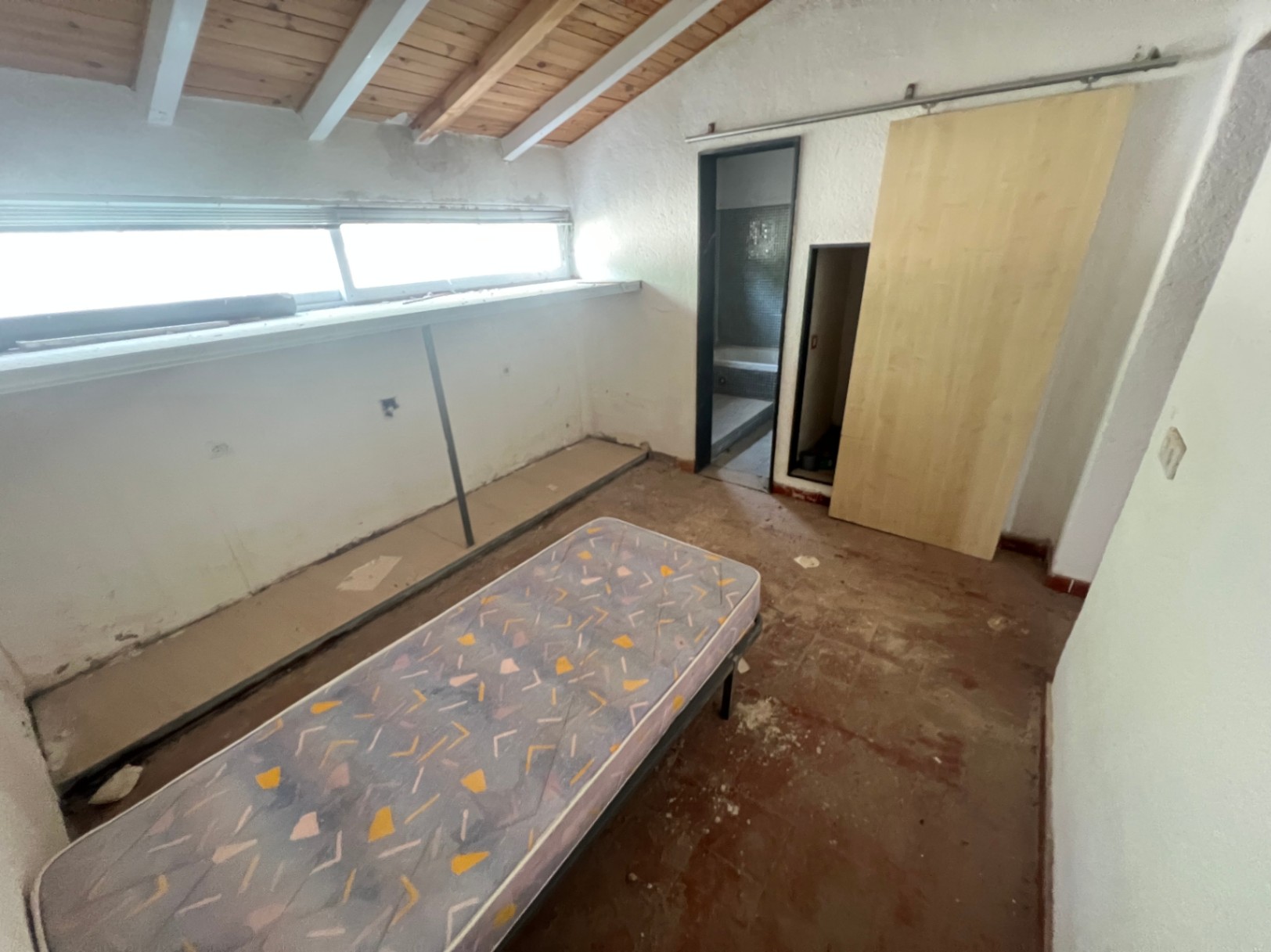 Schlafzimmer mit sichtbaren Holzbalken, langgestrecktem Fenster und angrenzendem Badezimmer. Ein renovierungsbedürftiger Raum mit viel Potenzial.