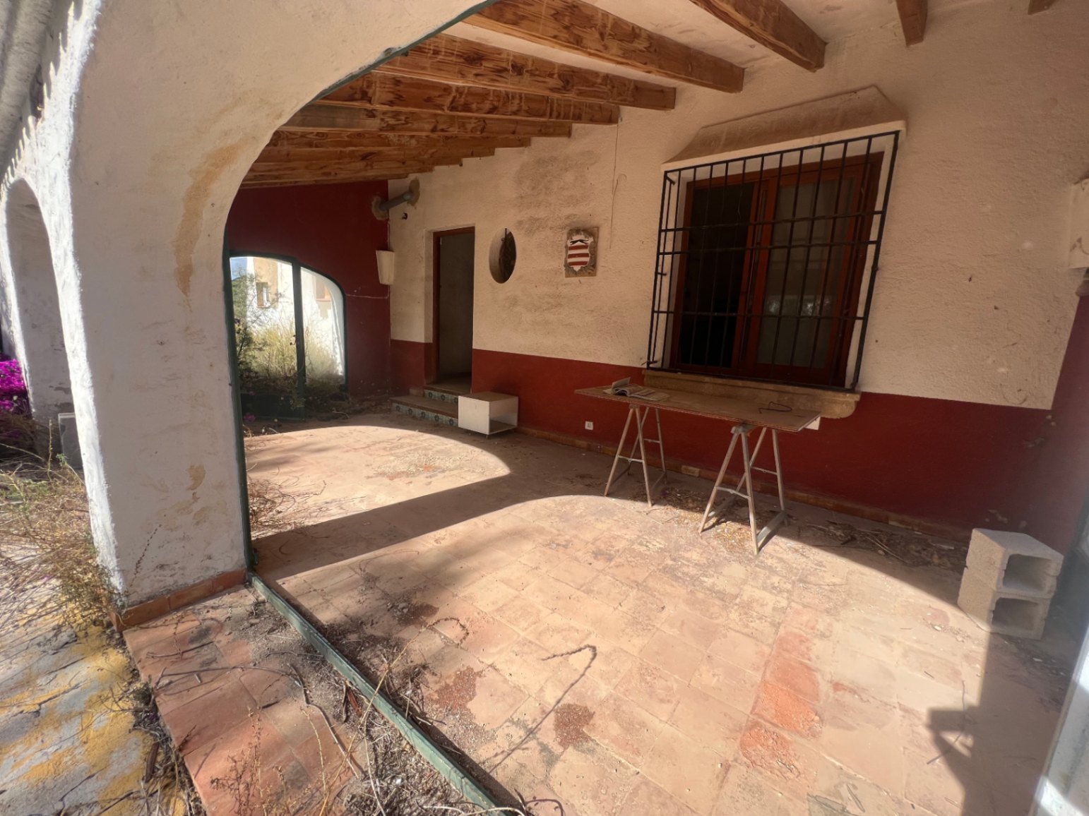 Traditionelle überdachte Terrasse mit sichtbaren Holzbalken, Terrakottaböden und mediterranen Rundbögen, die in den Garten führen.