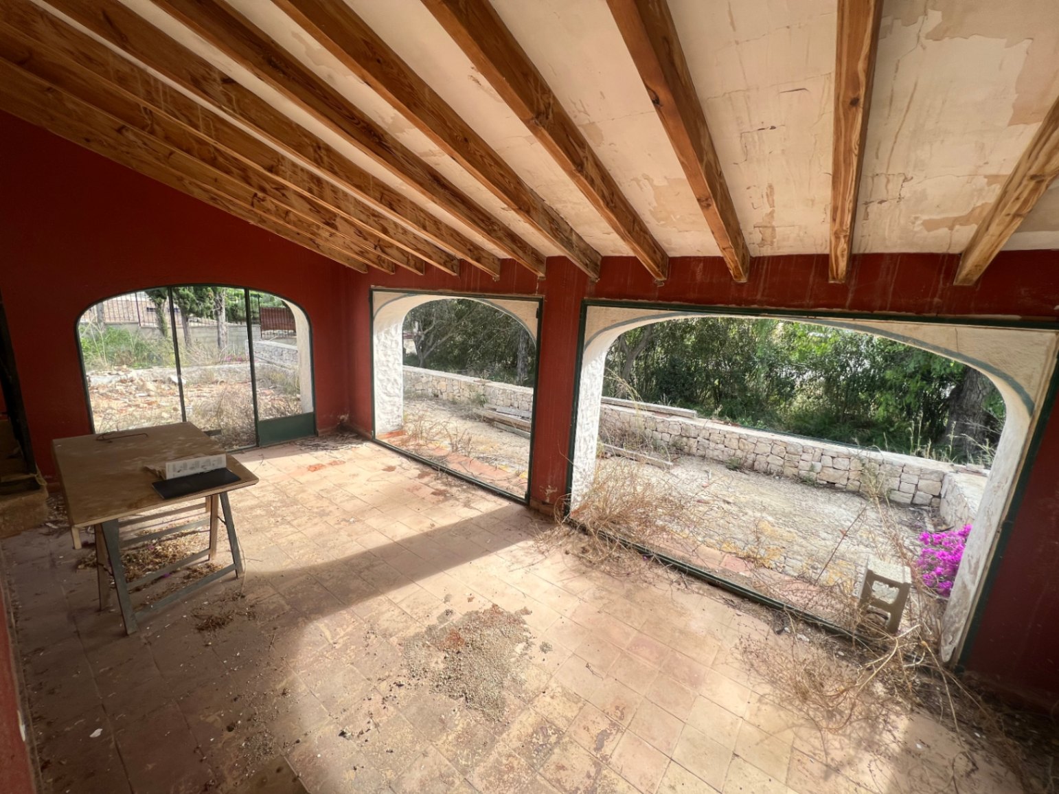 Überdachte Naya mit sichtbaren Holzbalken und Terrakottaboden. Große Rundbogenöffnungen bieten Ausblick auf den Garten und die ursprünglichen Steinmauern.