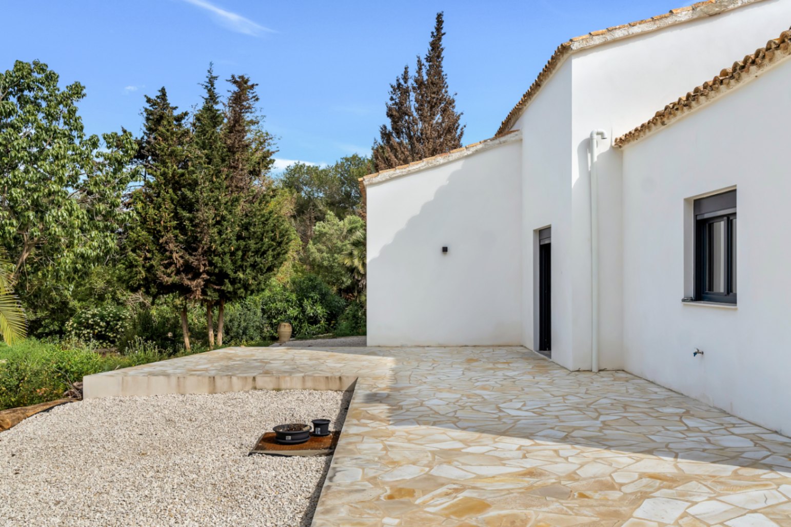 Außenansicht der Villa mit weißen Putzfassaden, Terrakottadach und einer großzügigen Natursteinterrasse inmitten mediterraner Bepflanzung.