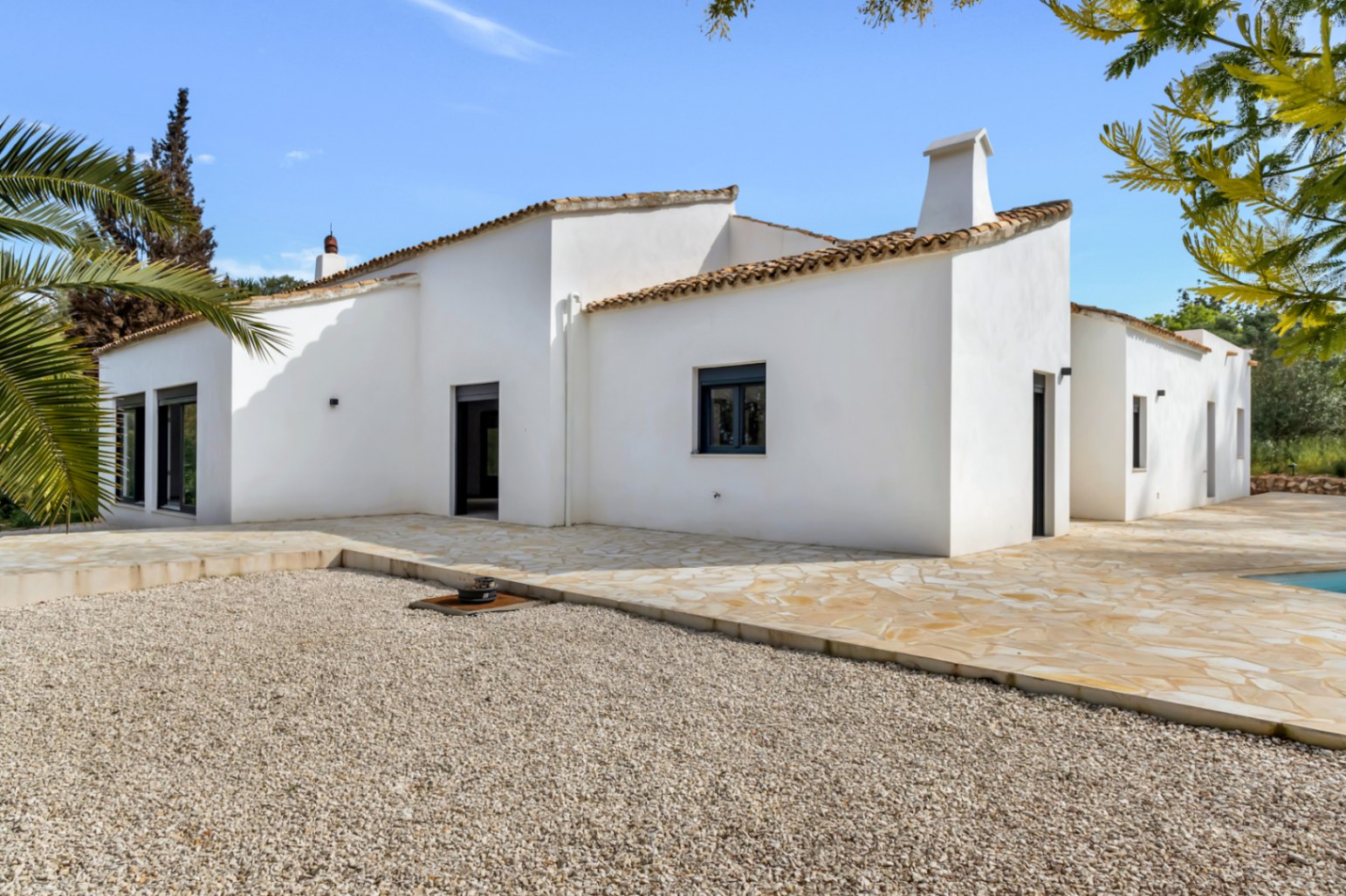 Außenansicht einer mediterranen Villa mit weißen Stuckwänden, Terrakotta-Dachziegeln und einer großen Natursteinterrasse neben einem Kiesbereich.