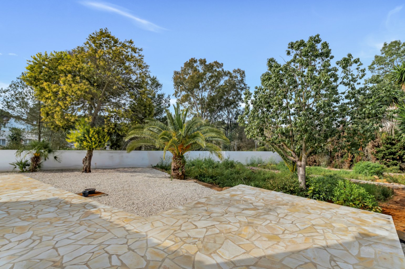 Außenbereich mit unregelmäßigen Natursteinplatten, angrenzend an einen mediterranen Garten mit Palmen, altem Baumbestand und gepflegten Kiesflächen.