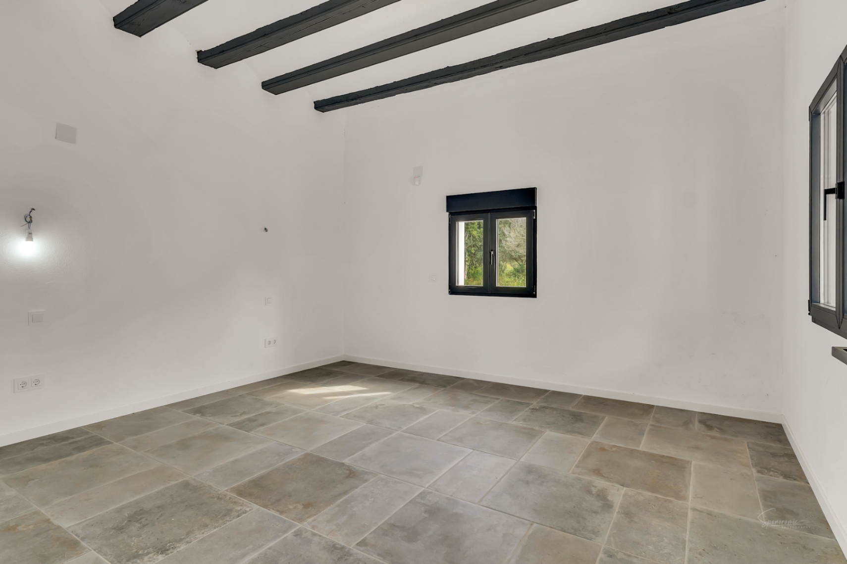 Heller Raum mit großformatigen Natursteinfliesen, hohen Decken mit dunklen Sichtholzbalken und schwarzen Fensterrahmen für natürliches Licht.