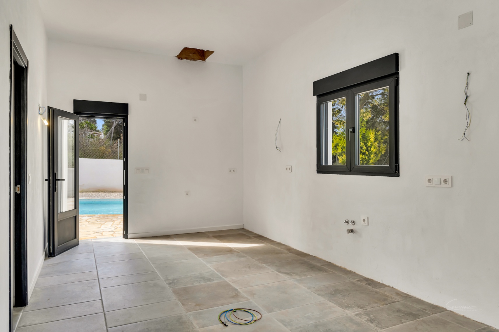 Innenraum in Renovierung mit Natursteinboden, schwarzen Fensterrahmen und Glastür, die direkt zum Pool und zur Außenterrasse führt.