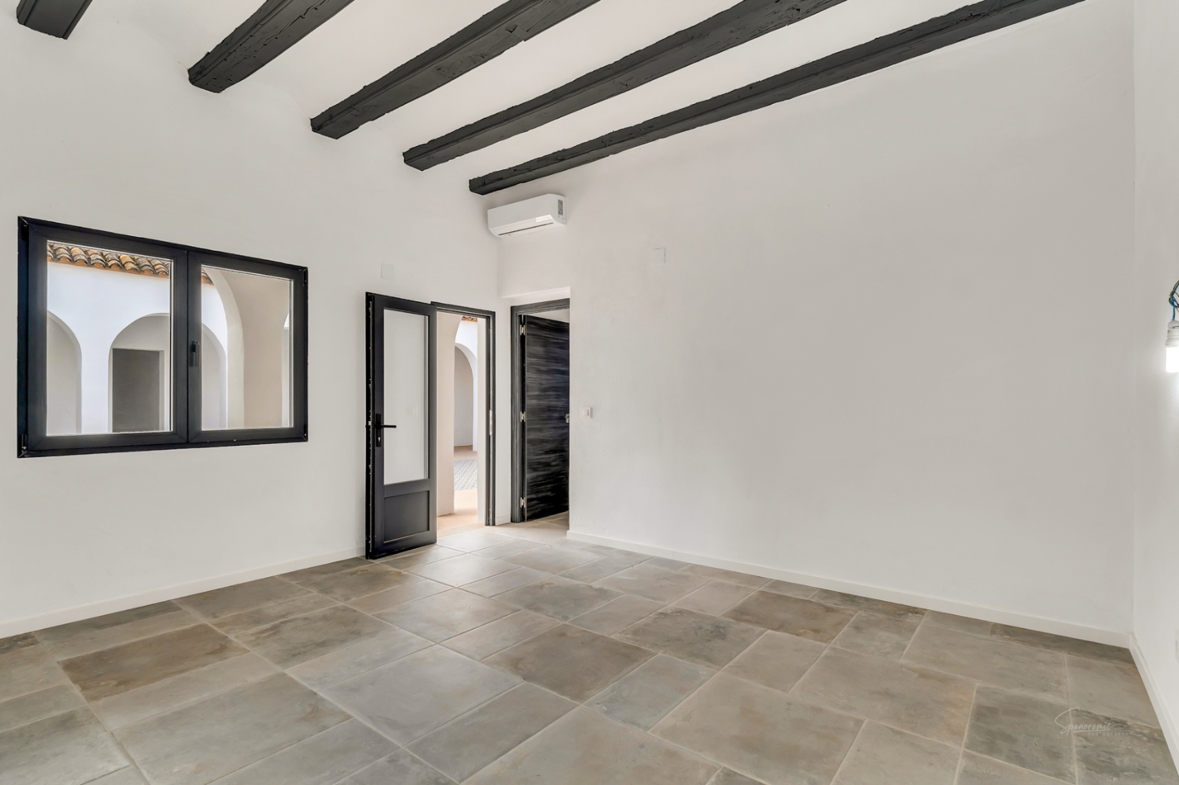 Innenraum mit dunklen Holzbalkendecken, Natursteinboden und Blick auf den bogenförmigen Innenhof durch schwarz gerahmte Fenster und Türen.