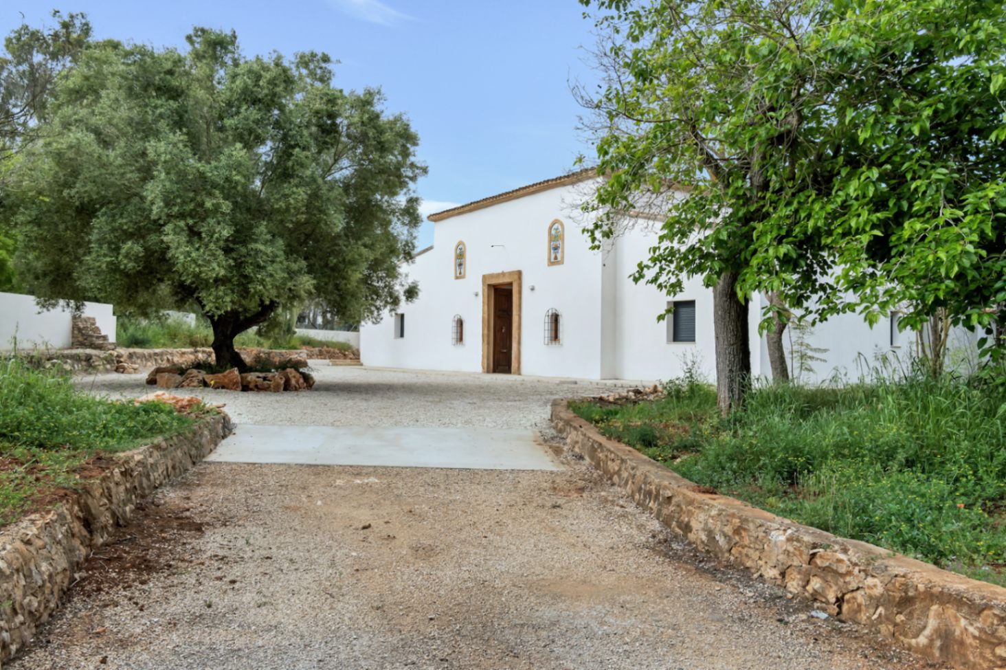 Mediterrane Fassade mit weißen Stuckwänden, Türrahmen aus Tosca-Stein und einer von Natursteinmauern und alten Olivenbäumen gesäumten Kieseinfahrt.
