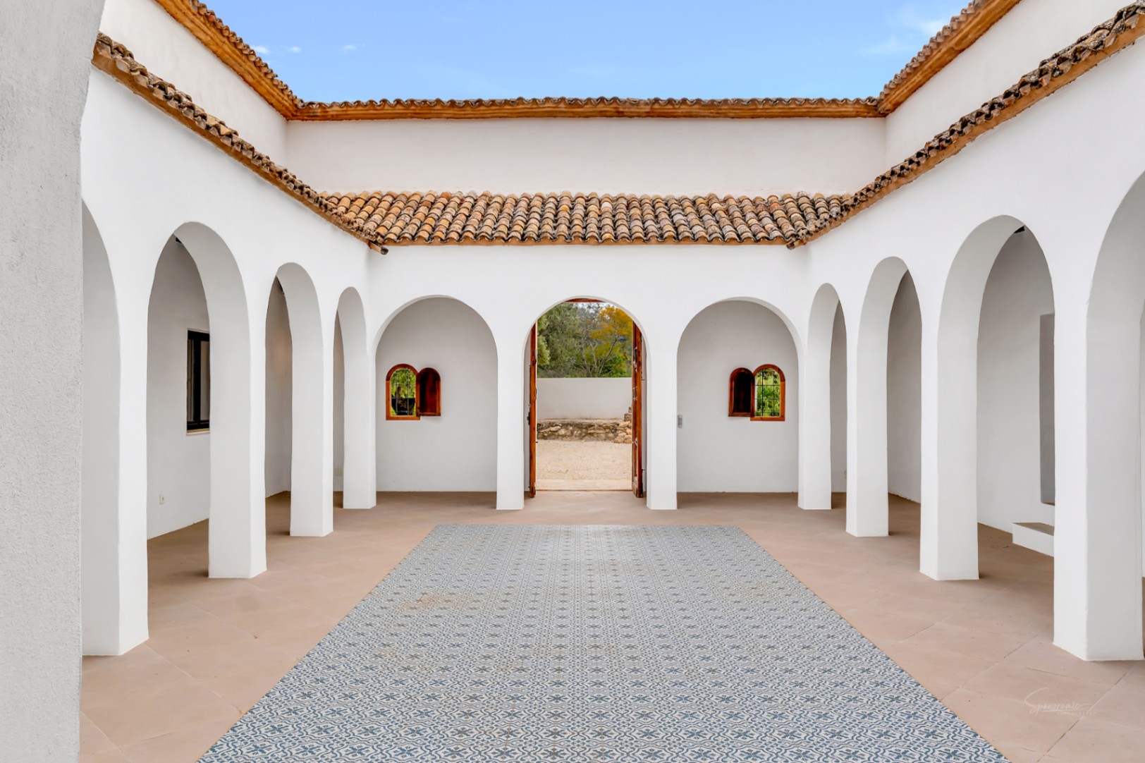 Mediterraner Innenhof mit weißen Rundbogen-Arkaden, Terrakotta-Dachziegeln und einem zentralen Bodenbereich mit dekorativen gemusterten Fliesen.