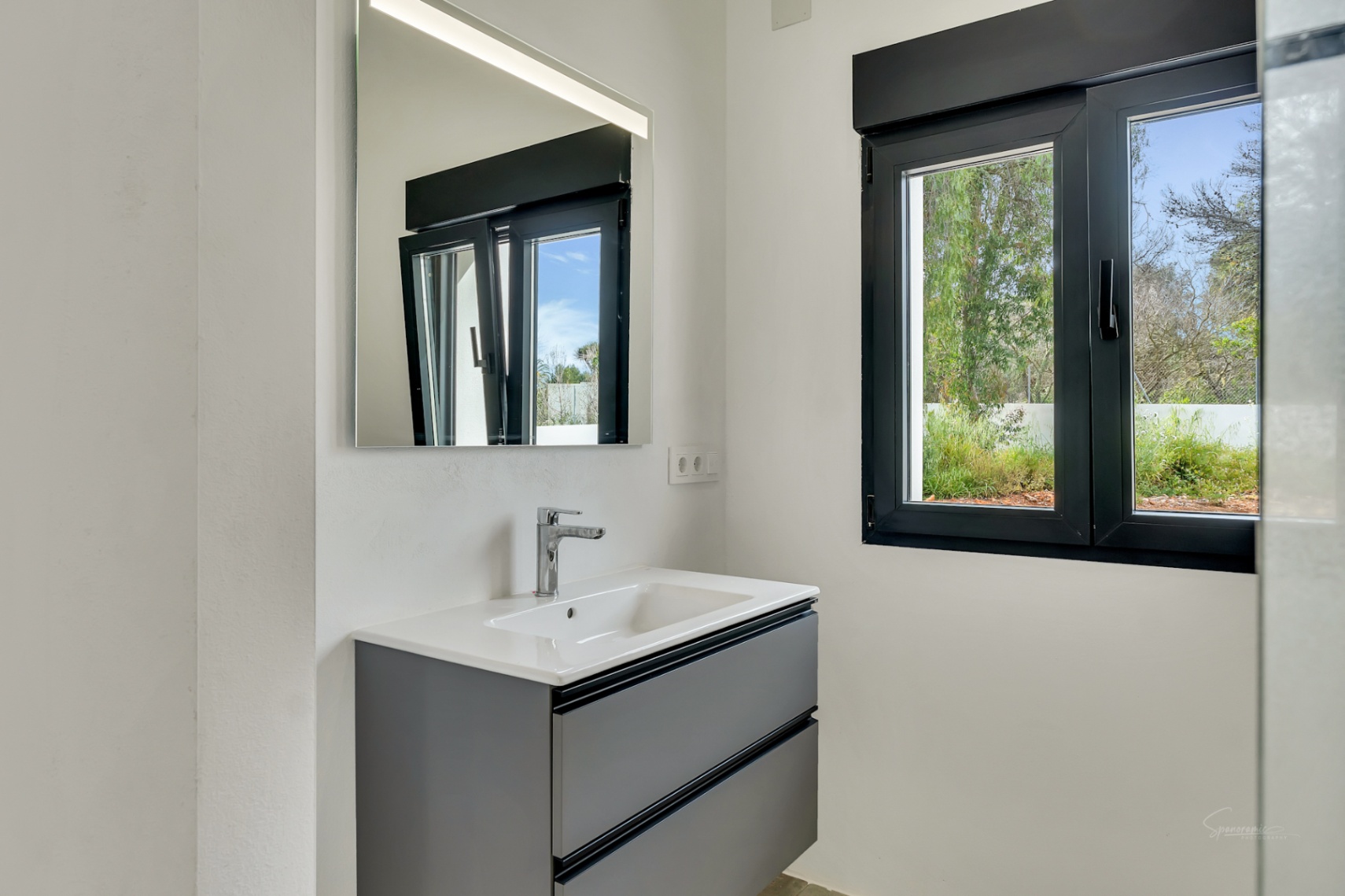 Modernes Badezimmer mit dunkelgrauem Waschtisch, beleuchtetem Spiegel und einem schwarzen Fensterrahmen, der Tageslicht und Gartenblick bietet.