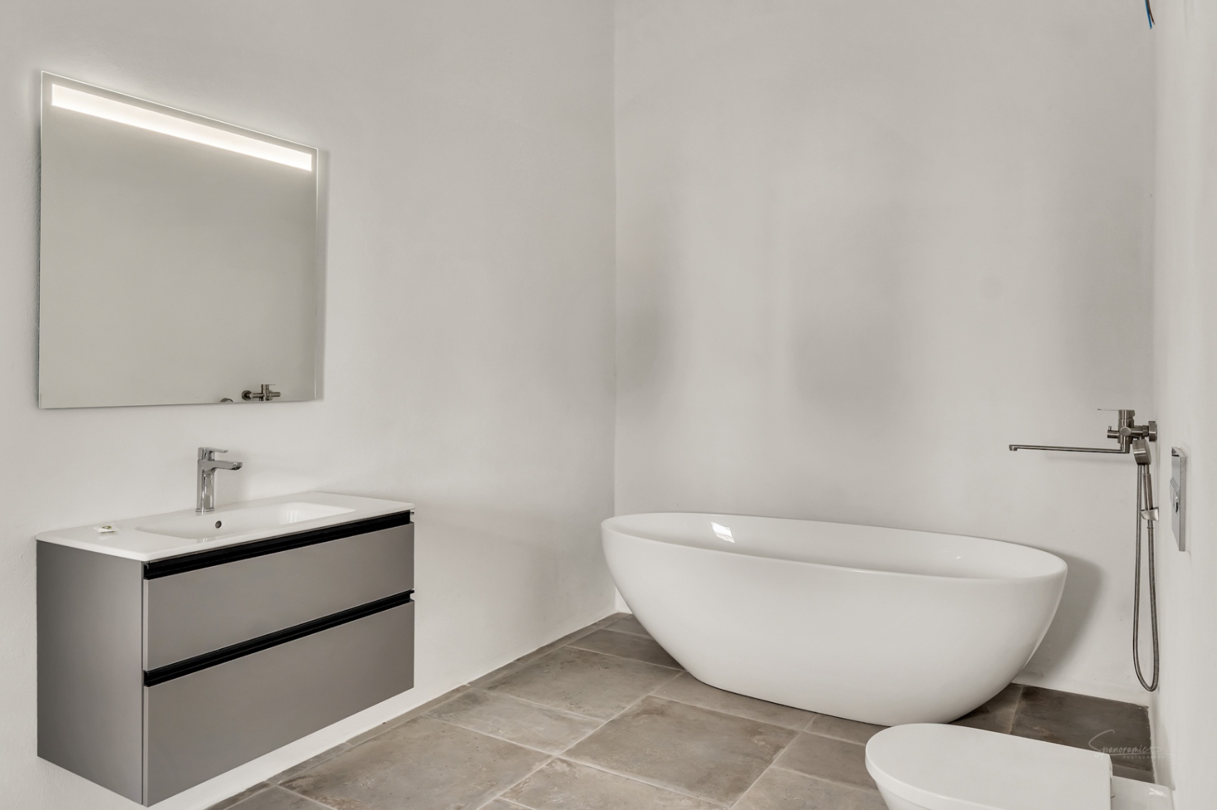 Modernes Badezimmer mit freistehender ovaler Badewanne, schwebendem grauem Waschtisch und hinterleuchtetem Spiegel auf Natursteinboden.