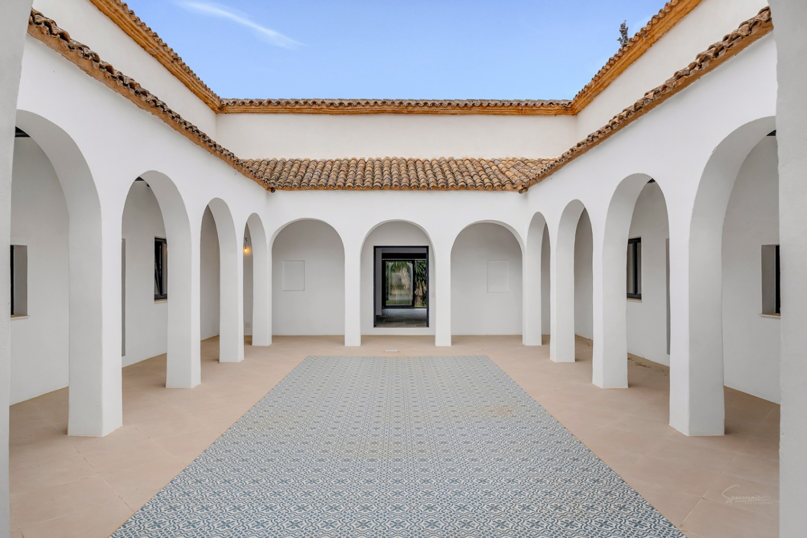 Zentraler mediterraner Innenhof mit symmetrischen weißen Rundbögen, traditionellen Terrakotta-Dachziegeln und dekorativen blau gemusterten Bodenfliesen.