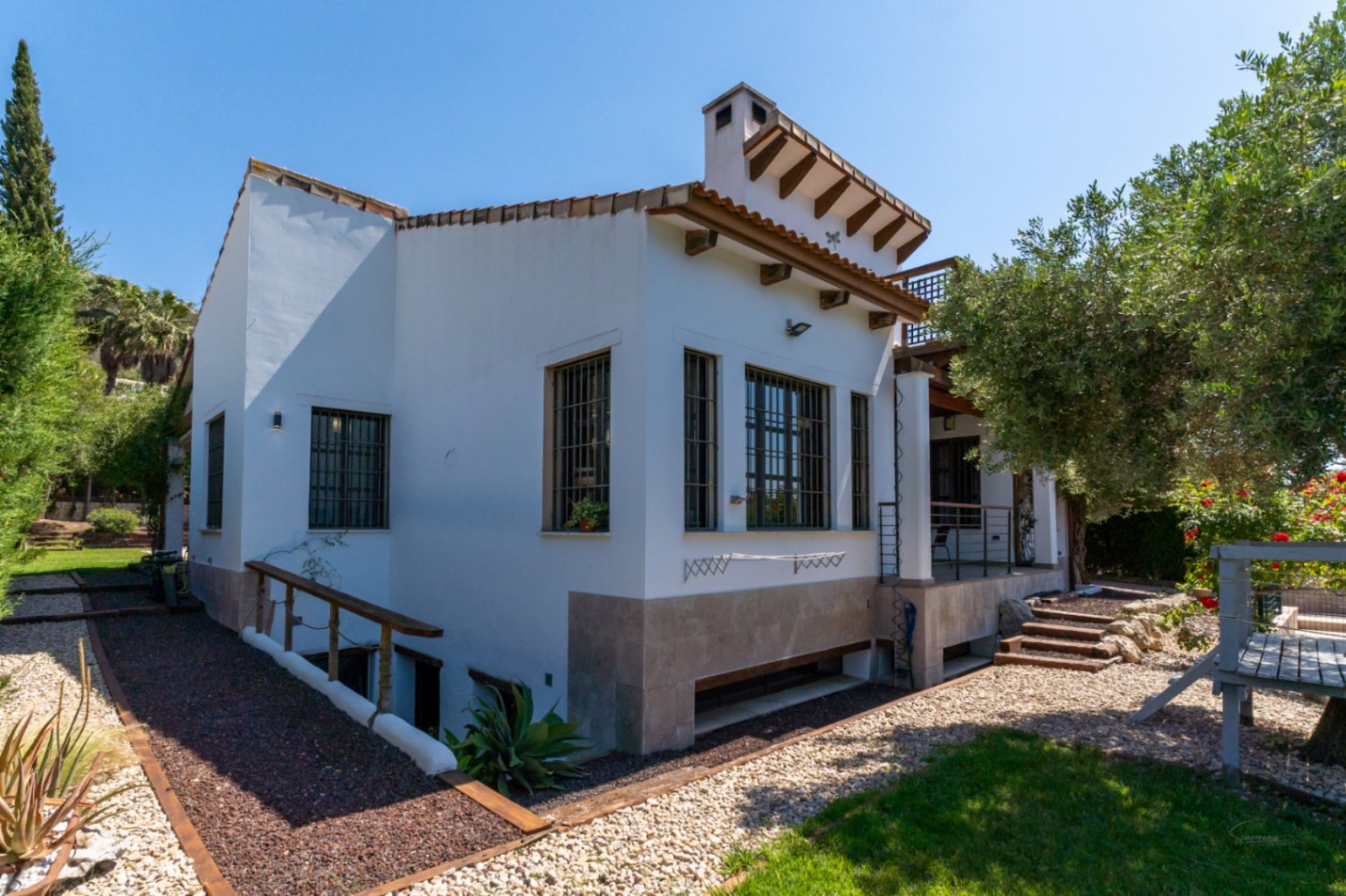 Außenansicht der Villa mit weißer Fassade, Holzbalken und Steinsockel. Der Garten bietet Kieswege und einen absteigenden Zugang zum Souterrain-Bereich.