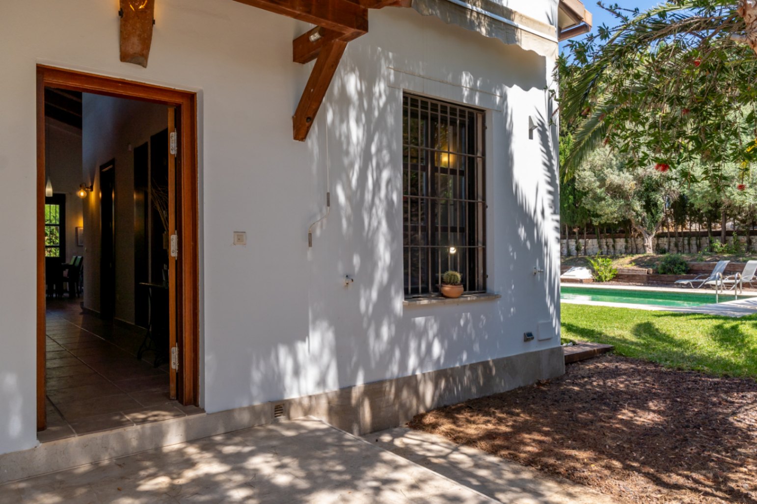 Außeneingang mit offener Holztür, weißer Fassade mit Palmenschatten und vergittertem Fenster mit Blick auf den gepflegten Garten und den Poolbereich.