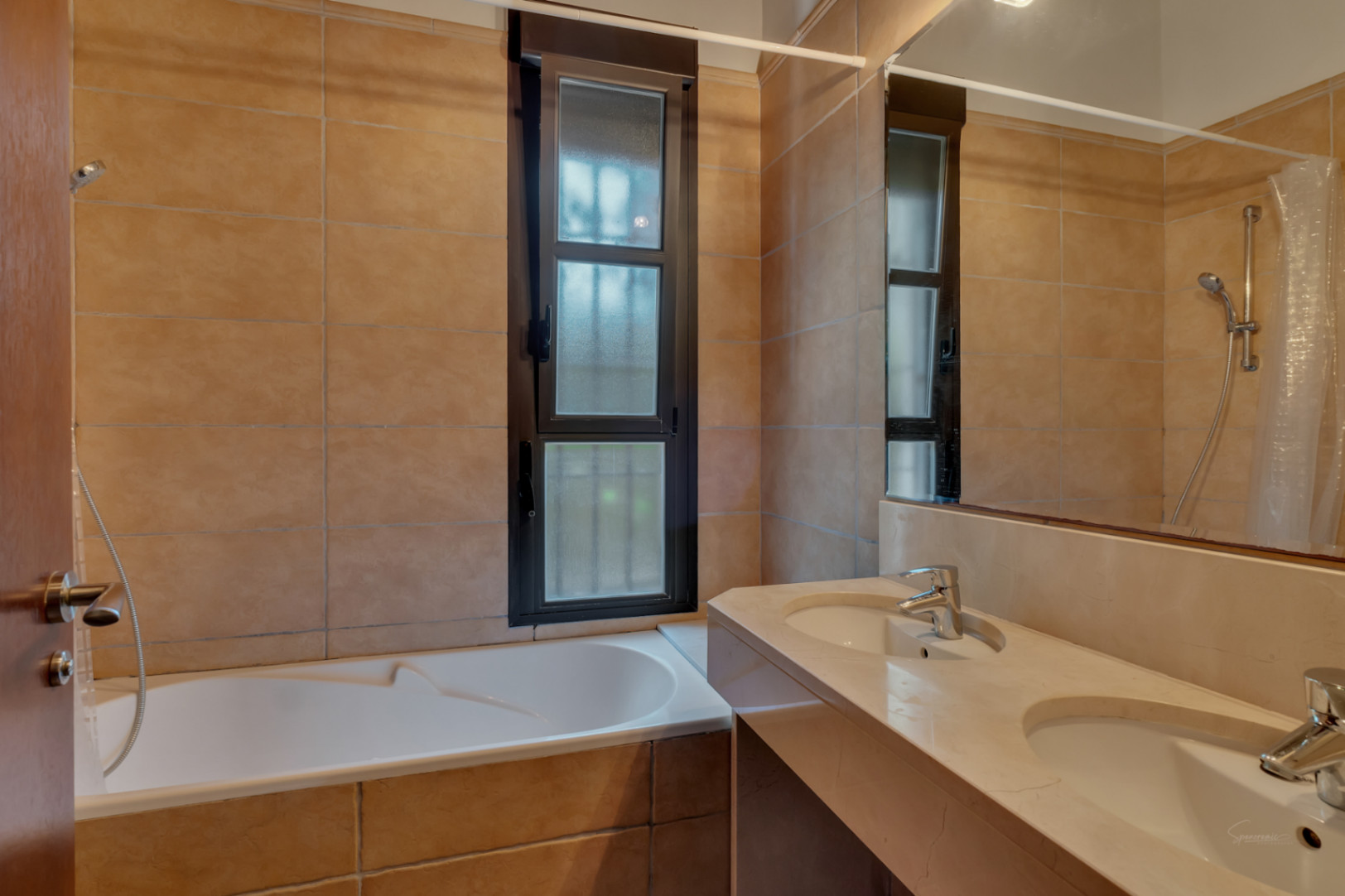 Badezimmer mit Doppelwaschtisch aus Stein, integrierter Badewanne und erdfarbenen Fliesen. Ein hohes Fenster mit Milchglas sorgt für natürliches Licht und Privatsphäre.