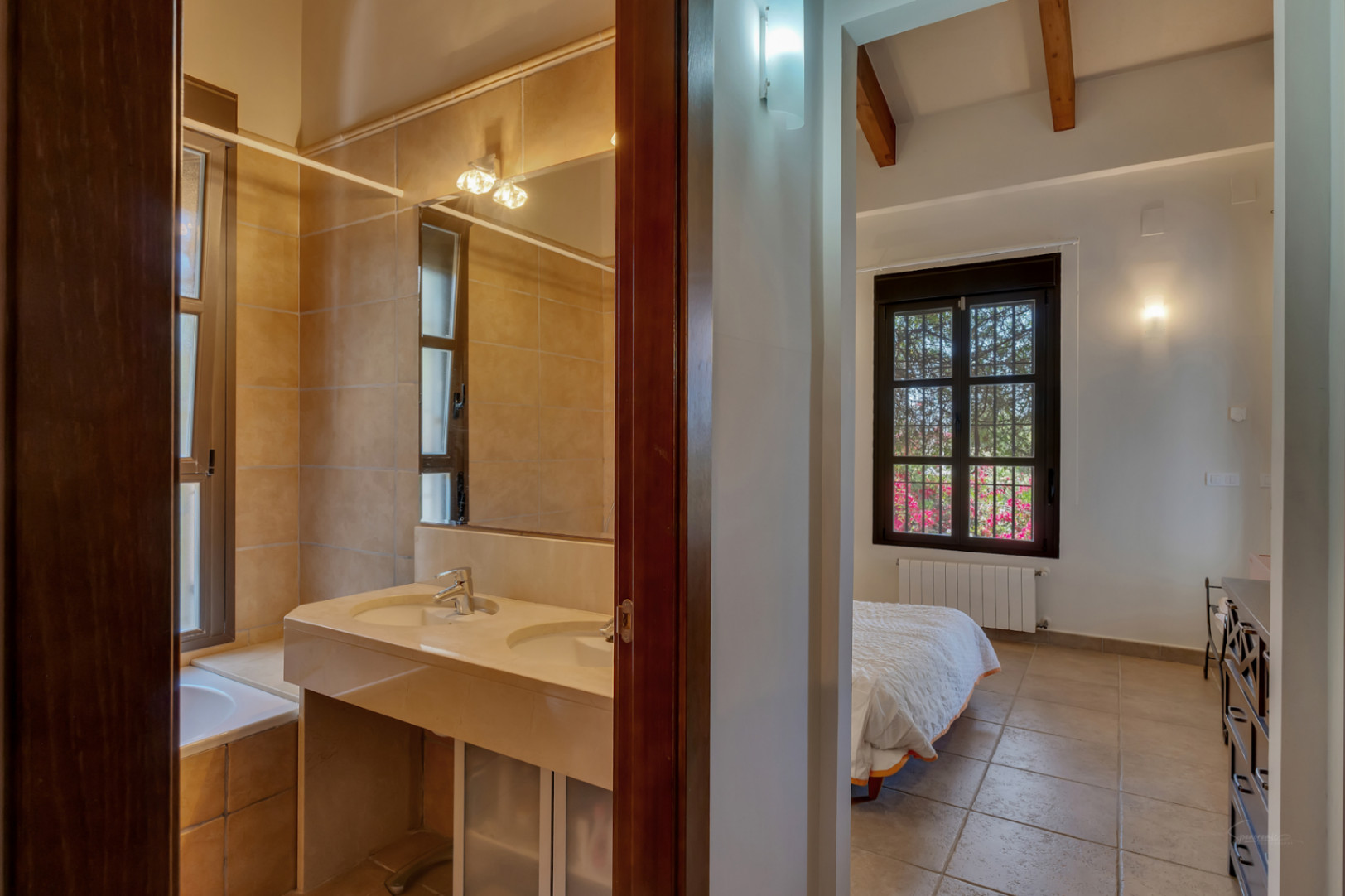 Blick vom Flur auf ein Badezimmer mit Doppelwaschbecken aus Stein und ein Schlafzimmer mit sichtbaren Holzbalken und Blick in den Garten.