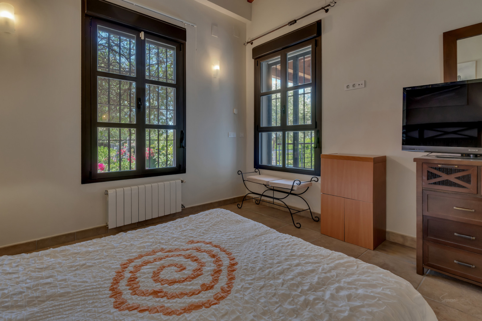 Helles Schlafzimmer mit dunklen Fensterrahmen, einem schmiedeeisernen Tischchen und Blick in den gepflegten Garten der Villa.