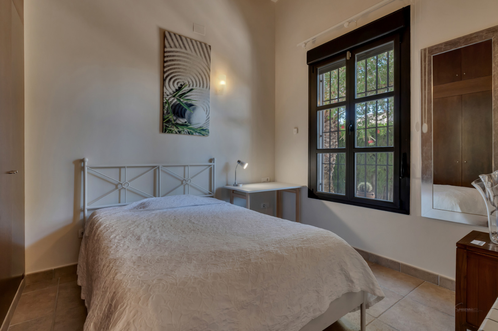 Helles Schlafzimmer mit weißem Metallbett, funktionalem Schreibtisch und dunklem Fensterrahmen mit Blick auf den mediterranen Garten.