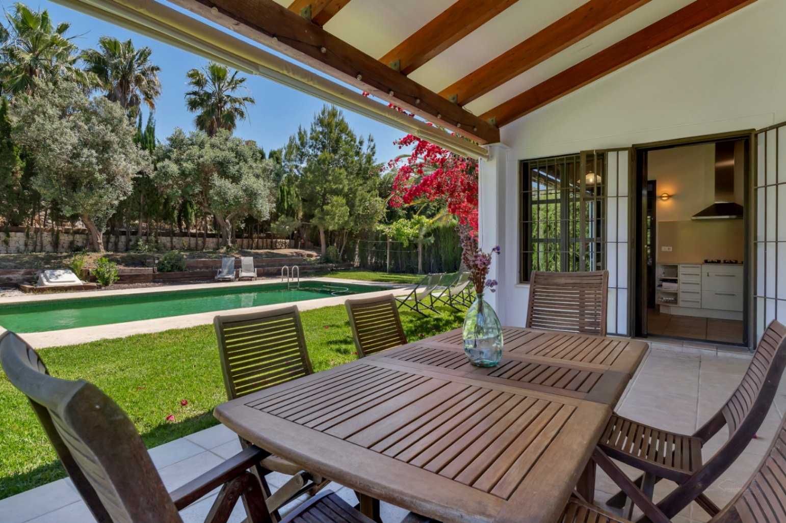 Überdachte Terrasse mit Holzbalken und Essbereich im Freien, mit Blick auf den mediterranen Garten und den rechteckigen Pool.