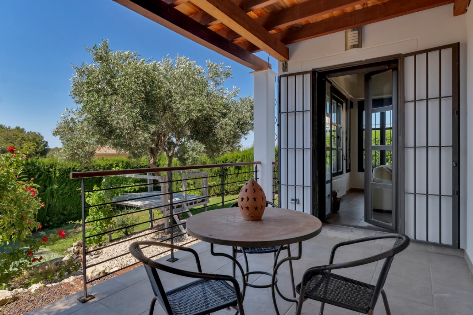 Überdachte Terrasse mit Holzbalkendecke, Fliesenboden und Schmiedeeisenmöbeln mit Blick auf den mediterranen Garten und einen Olivenbaum.