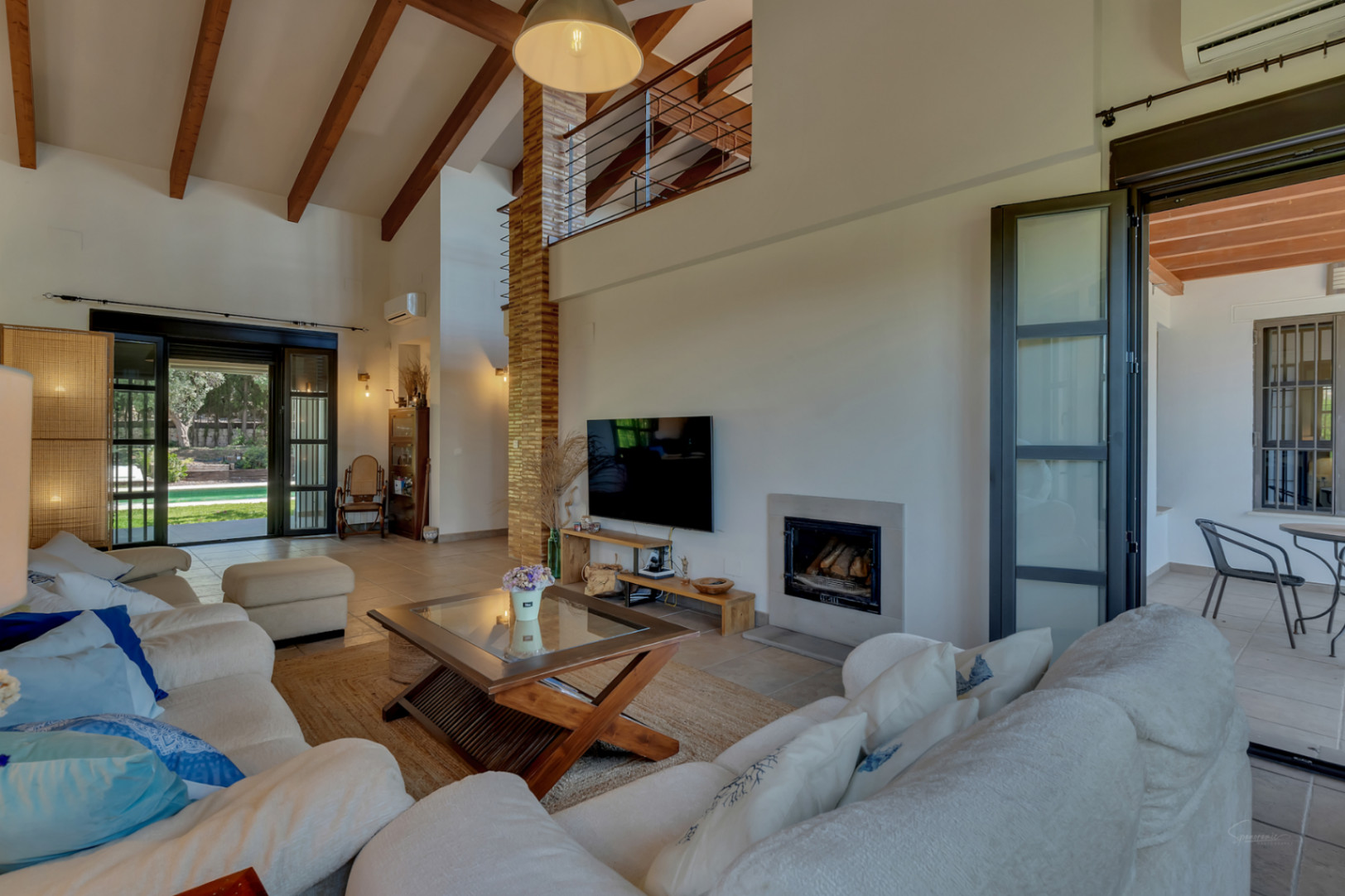 Wohnzimmer mit hohen Decken und Holzbalken, integriertem Kamin und direktem Zugang zur Terrasse und zum Garten durch große Glastüren.