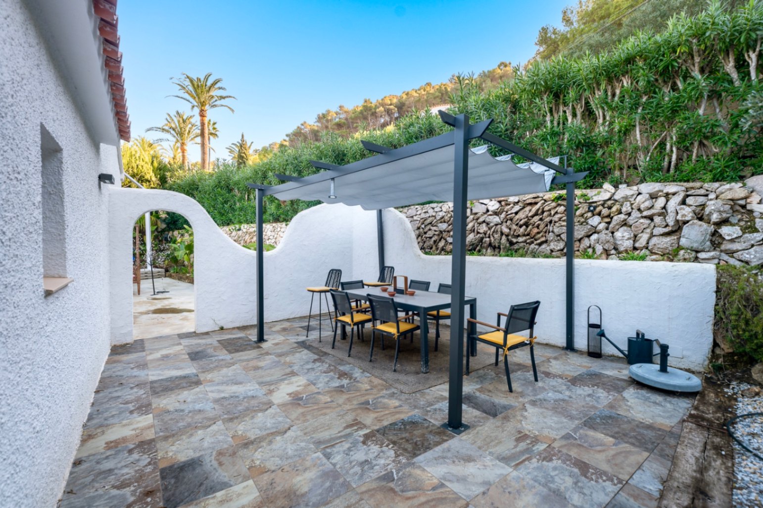 Außen-Essbereich mit moderner Pergola, Natursteinboden und traditionellen weißen Mauern vor einer begrünten Hügellandschaft.