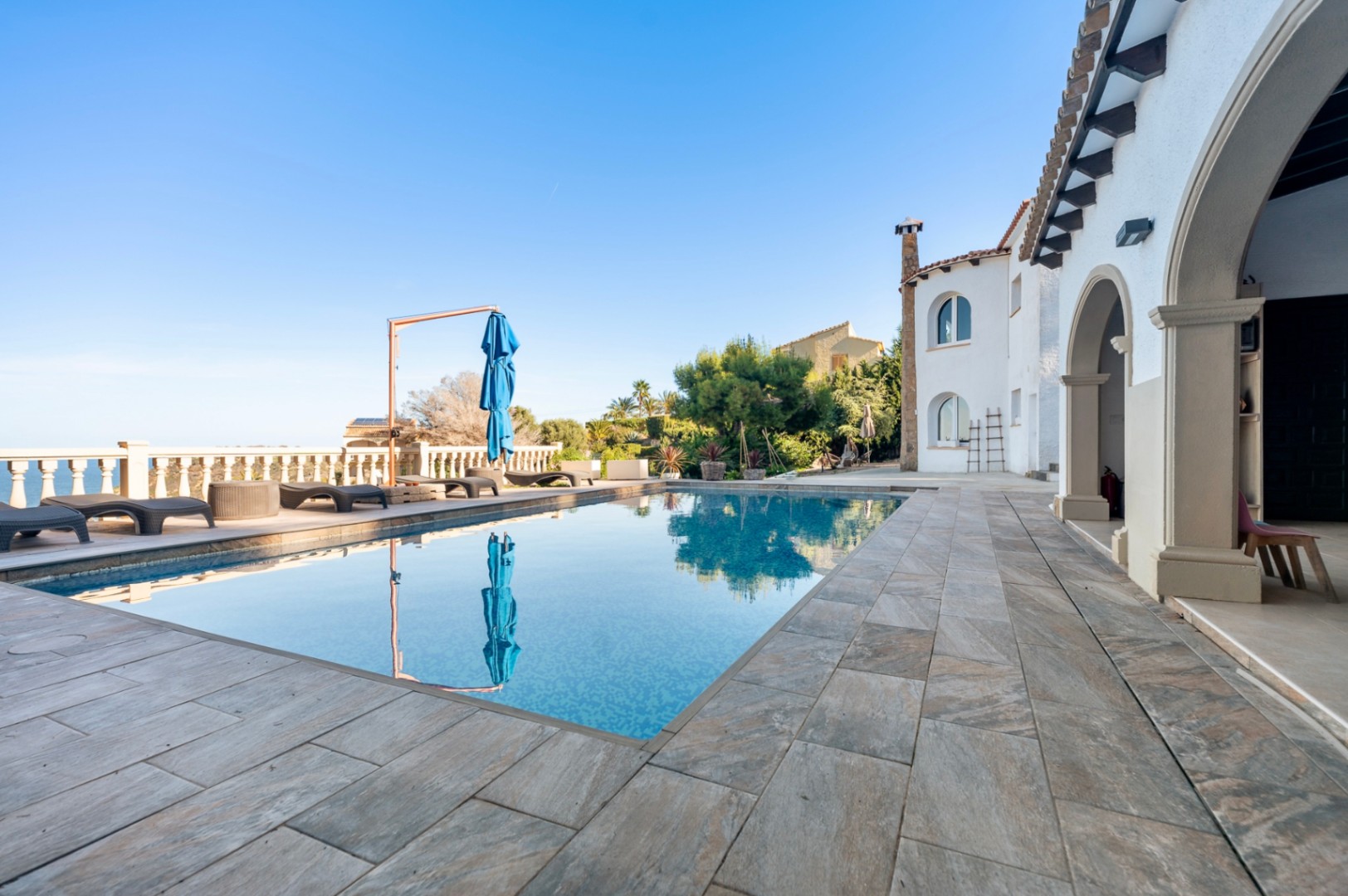 Außen-Terrasse mit rechteckigem Pool, Natursteinpflaster und Sonnenliegen mit Blick auf das Meer und die traditionelle Architektur der Villa mit Rundbögen.
