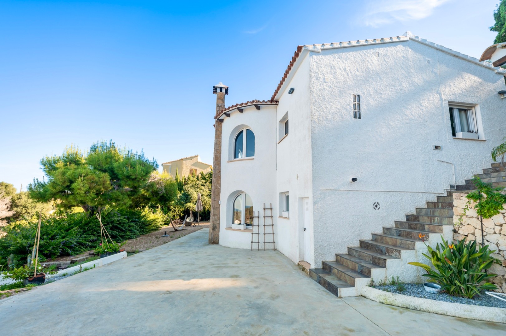 Außenansicht der Villa mit weiß verputzten Wänden, Rundbogenfenstern und einer Steintreppe zum Obergeschoss, eingebettet in einen Garten mit Pinien und mediterranen Pflanzen.
