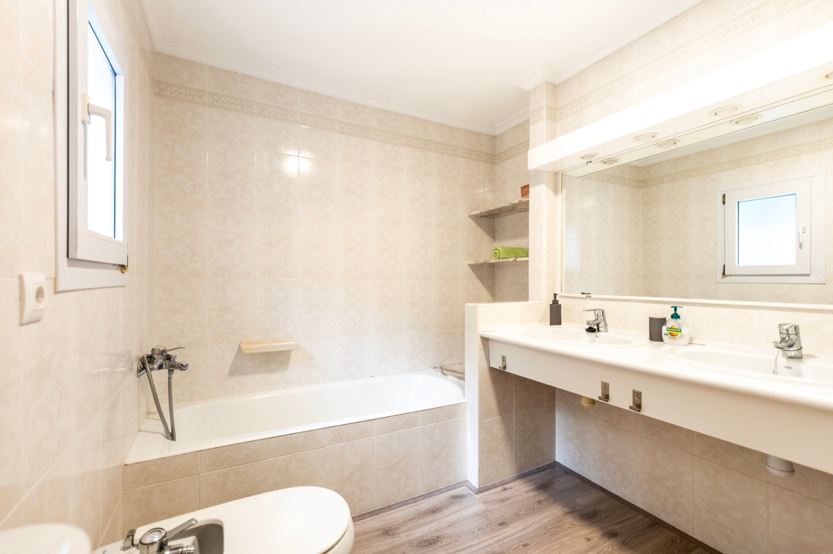 Badezimmer mit Badewanne, bidet und Doppelwaschtisch. Ausgestattet mit cremefarbenen Fliesen, einem großen beleuchteten Spiegel und Bodenbelag in Holzoptik.