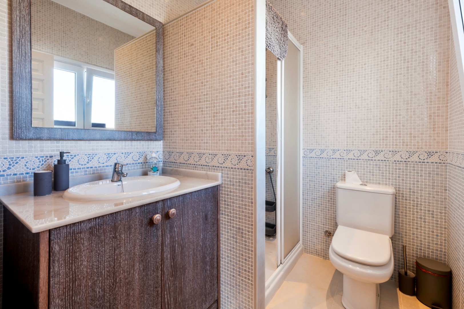 Badezimmer mit beigen Mosaikfliesen, blauer Wellenbordüre, dunklem Holzwaschtisch mit Marmorplatte und integrierter Duschkabine.