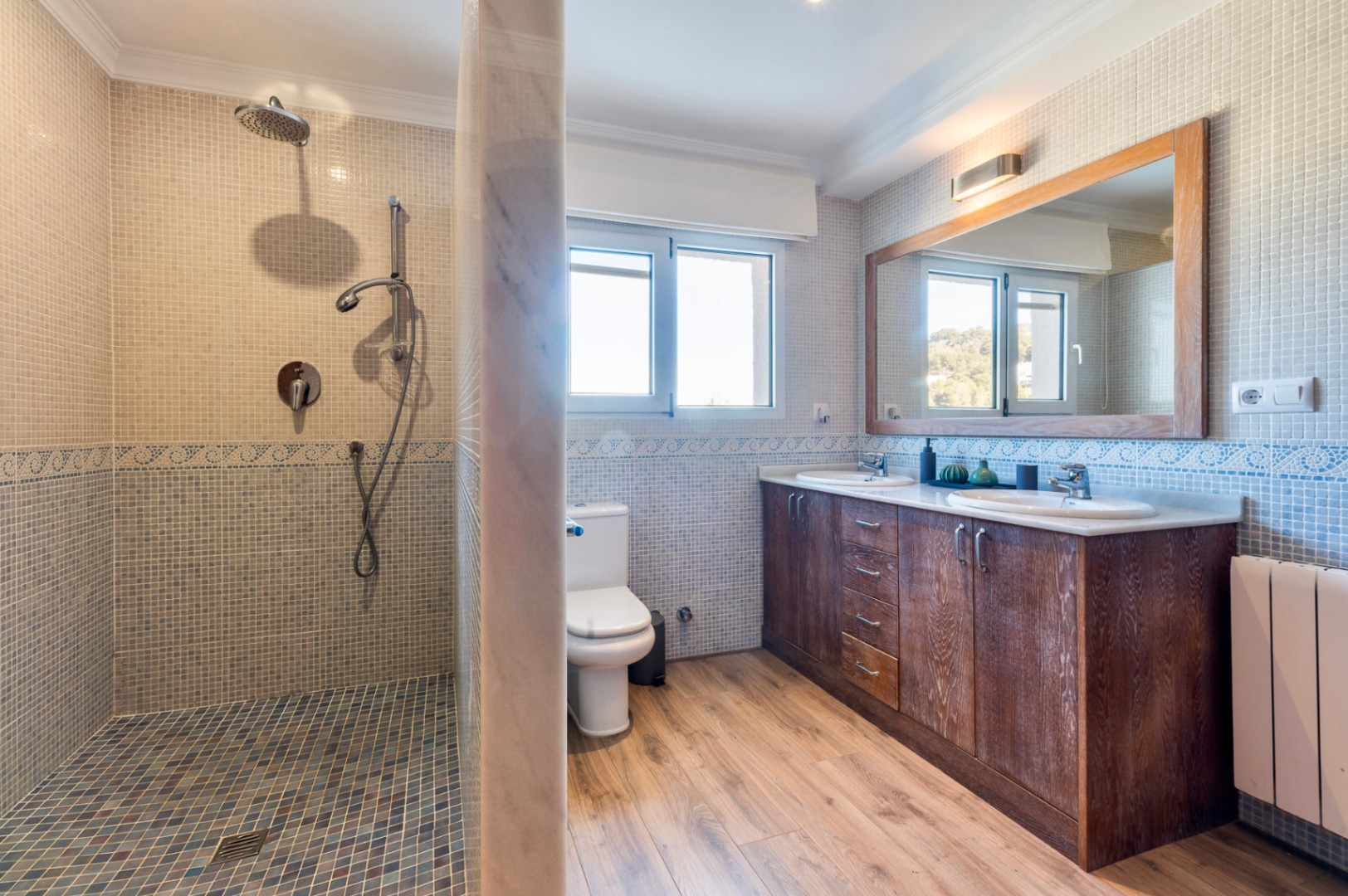 Badezimmer mit Mosaik-Dusche, Doppelwaschtisch aus Holz und Boden in Holzoptik. Ein Fenster sorgt für natürliches Licht und Belüftung.