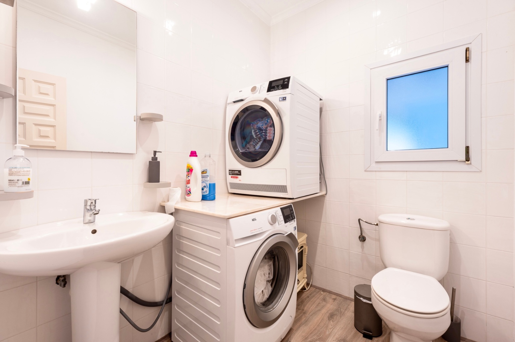 Funktionaler Wasch- und Toilettenraum mit weiß gefliesten Wänden, Standwaschbecken und einer AEG Waschmaschinen-Trockner-Kombination auf Laminatboden.