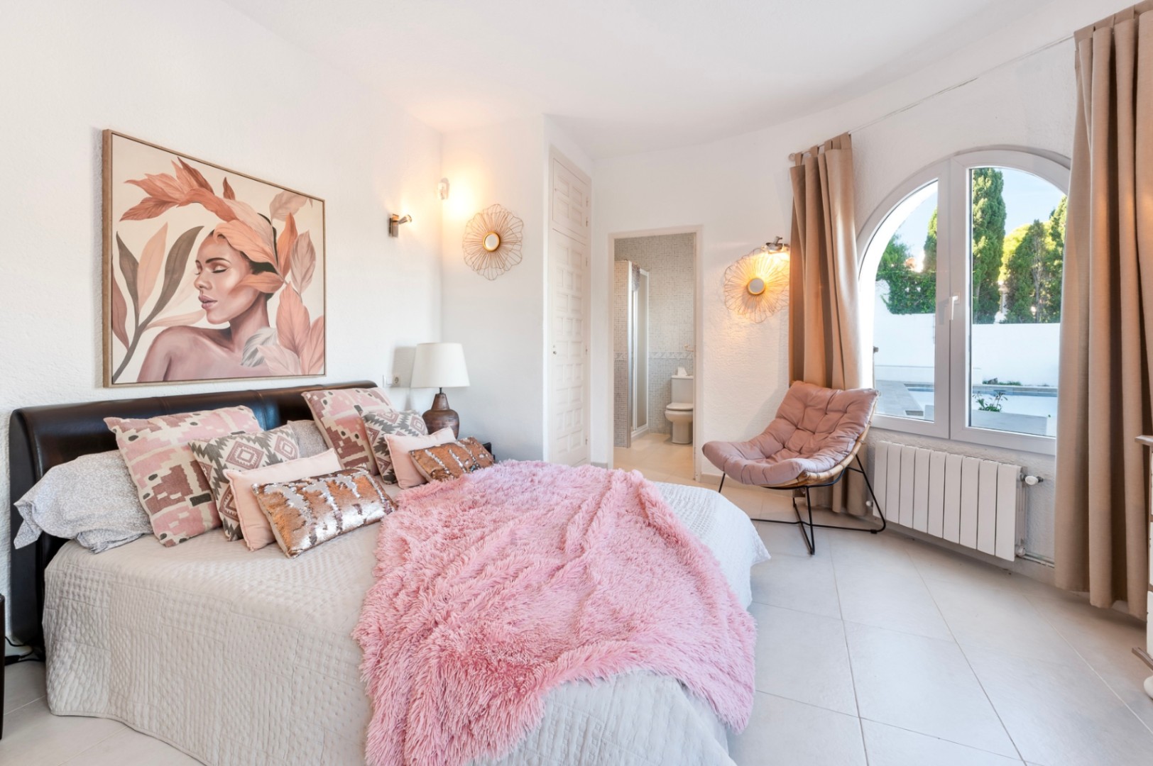 Helles Schlafzimmer mit En-Suite-Badezimmer, Rundbogenfenster mit Blick nach draußen und moderner Dekoration in Rosatönen auf hellem Fliesenboden.