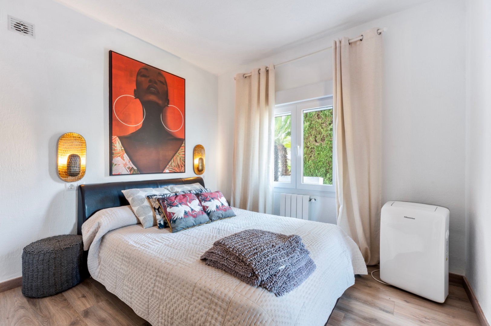 Helles Schlafzimmer mit Laminatboden, Doppelbett mit dunklem Kopfteil und goldenen Wandleuchten. Ausgestattet mit mobilem Klimagerät und Blick ins Grüne durch das Fenster.